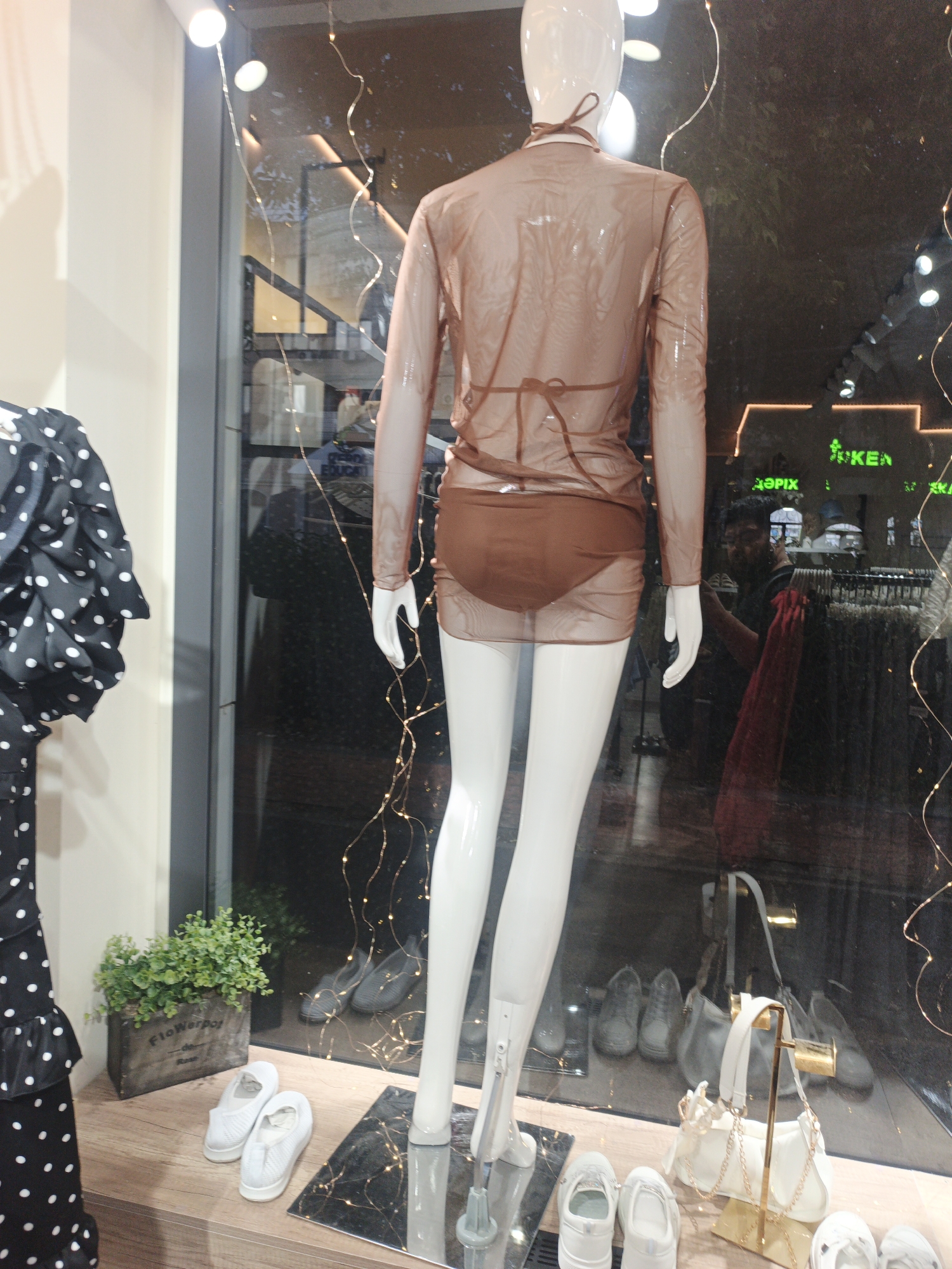 A full-body mannequin in a sheer, brown, long-sleeved dress is displayed in a shop window. The mannequin is positioned to the right of a black dress with white polka dots. In the foreground, to the left of the mannequin's legs, are two white flat shoes and a wooden planter with a green plant. In the background, visible through the shop window, are shelves filled with clothing, and a sign that reads "IKEA" in green lettering. The reflection in the window shows several pairs of shoes and handbags on display. The scene appears to be a retail store in Shymkent, Kazakhstan.