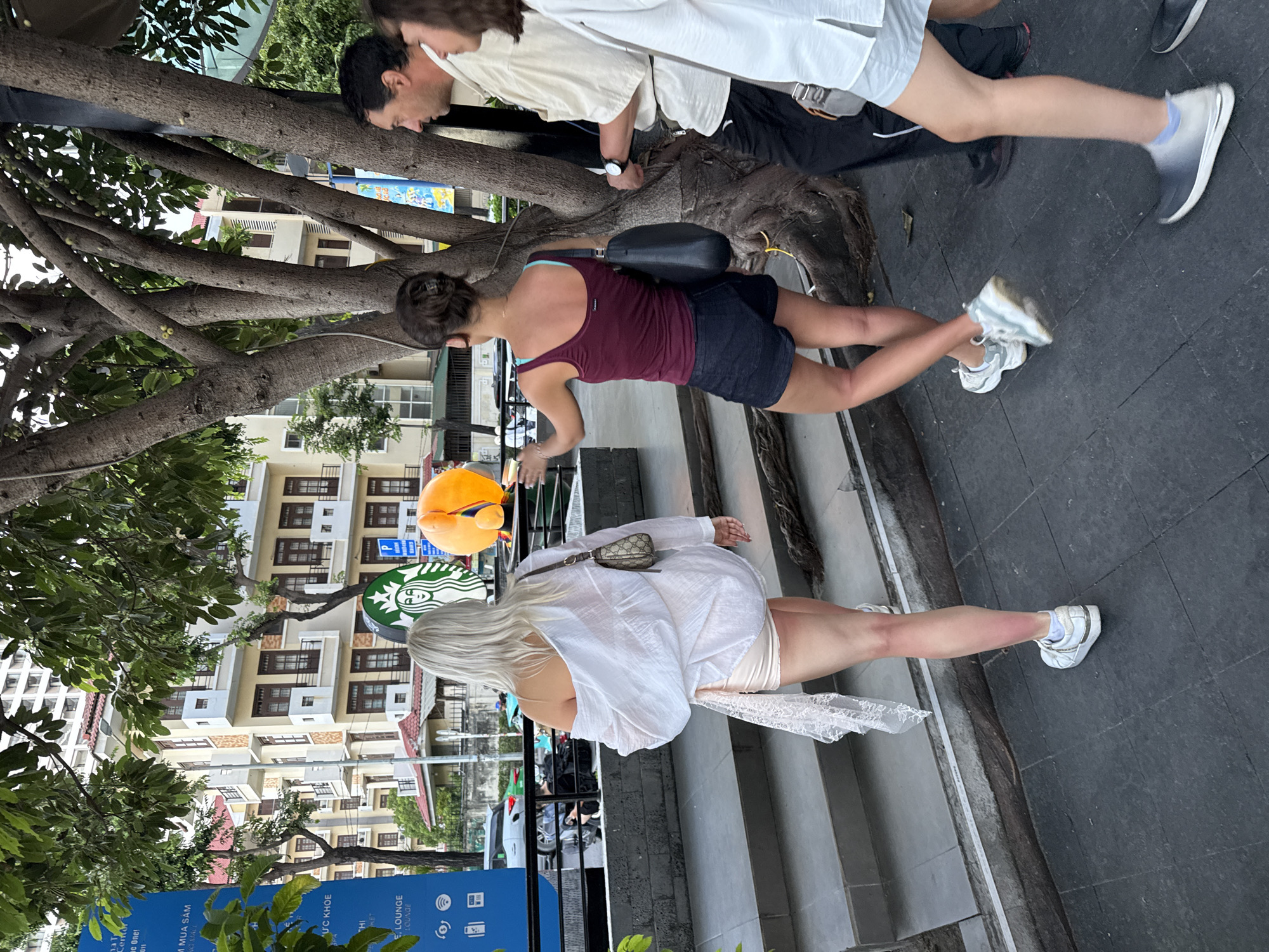 This is a medium shot taken from a slightly low angle, showing a street scene in Nha Trang Ward, Vietnam. In the foreground, a woman with platinum blonde hair walks away from the camera, wearing an oversized white shirt over a light-colored skirt and white sneakers. A small designer purse is slung across her shoulder. To her right, another woman, viewed from the back, wears a dark red tank top and dark shorts, with a black purse. She appears to be gesturing with her left hand towards a bright orange object, possibly a balloon, held by someone out of frame. Behind them, to the left, a blue sign with white text and icons is partially visible. A Starbucks logo is also present in the background, to the right of the blonde woman. Further back, a multi-story building with beige walls and dark windows is visible, along with trees and a street with cars. The ground in the foreground is paved with dark tiles, and there are concrete steps leading up to a raised area. The overall lighting suggests it is daytime.