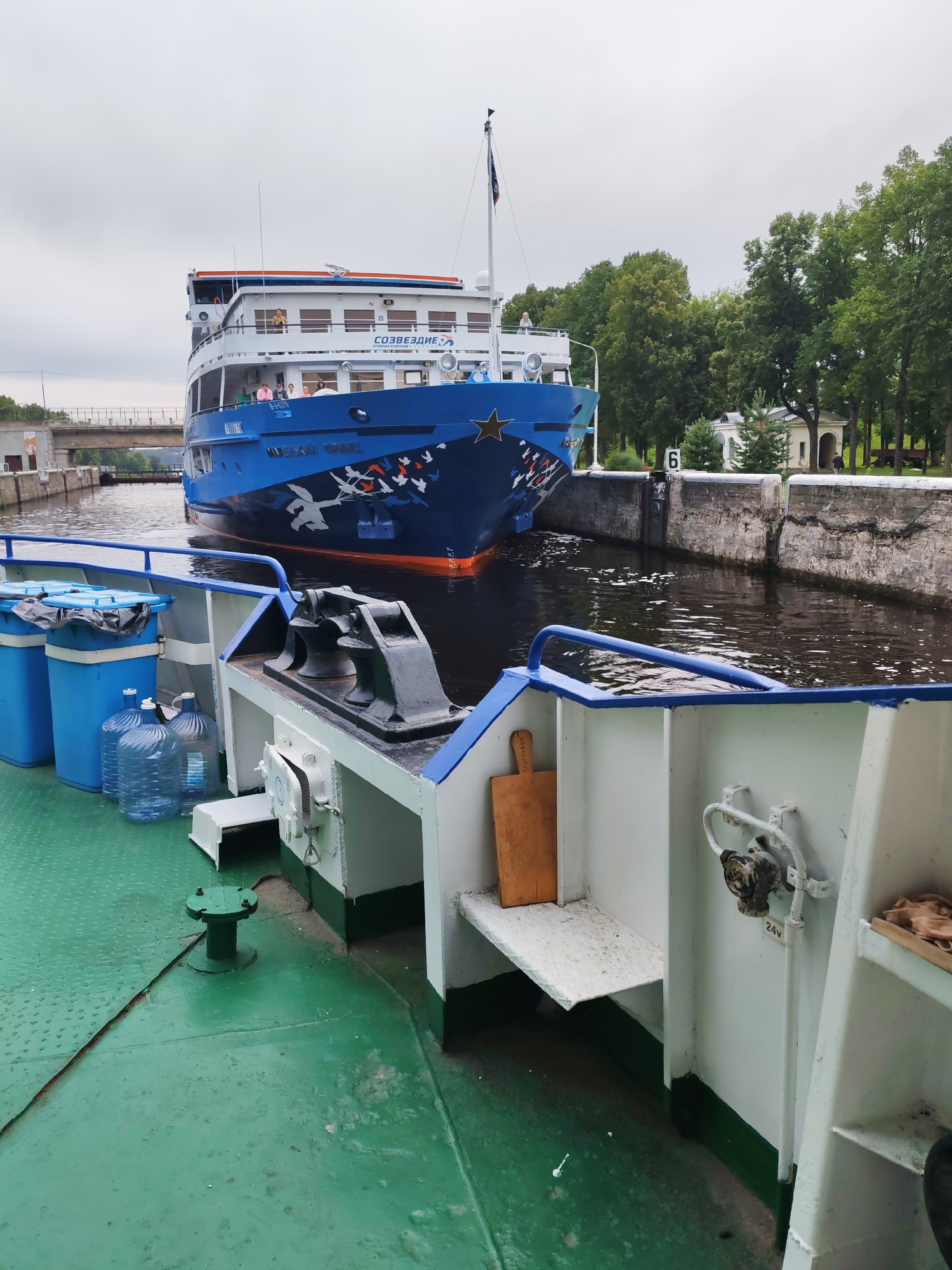 In the city of Vasil'ki, Russia, a blue and white passenger boat with "CO3BE3ДИE" and "WO3CHCKNI TPAH3" written on it is navigating through a canal lock. The boat is decorated with a large star and silhouettes of birds. On the deck of the passenger boat, several individuals are visible. In the foreground, another vessel with a green deck is present, featuring blue bins, large water bottles, and a black mooring bitt. The canal walls are constructed from stone, and a metal gate is partially visible. Trees line the banks of the canal, and a bridge can be seen in the distance. The sky is overcast.