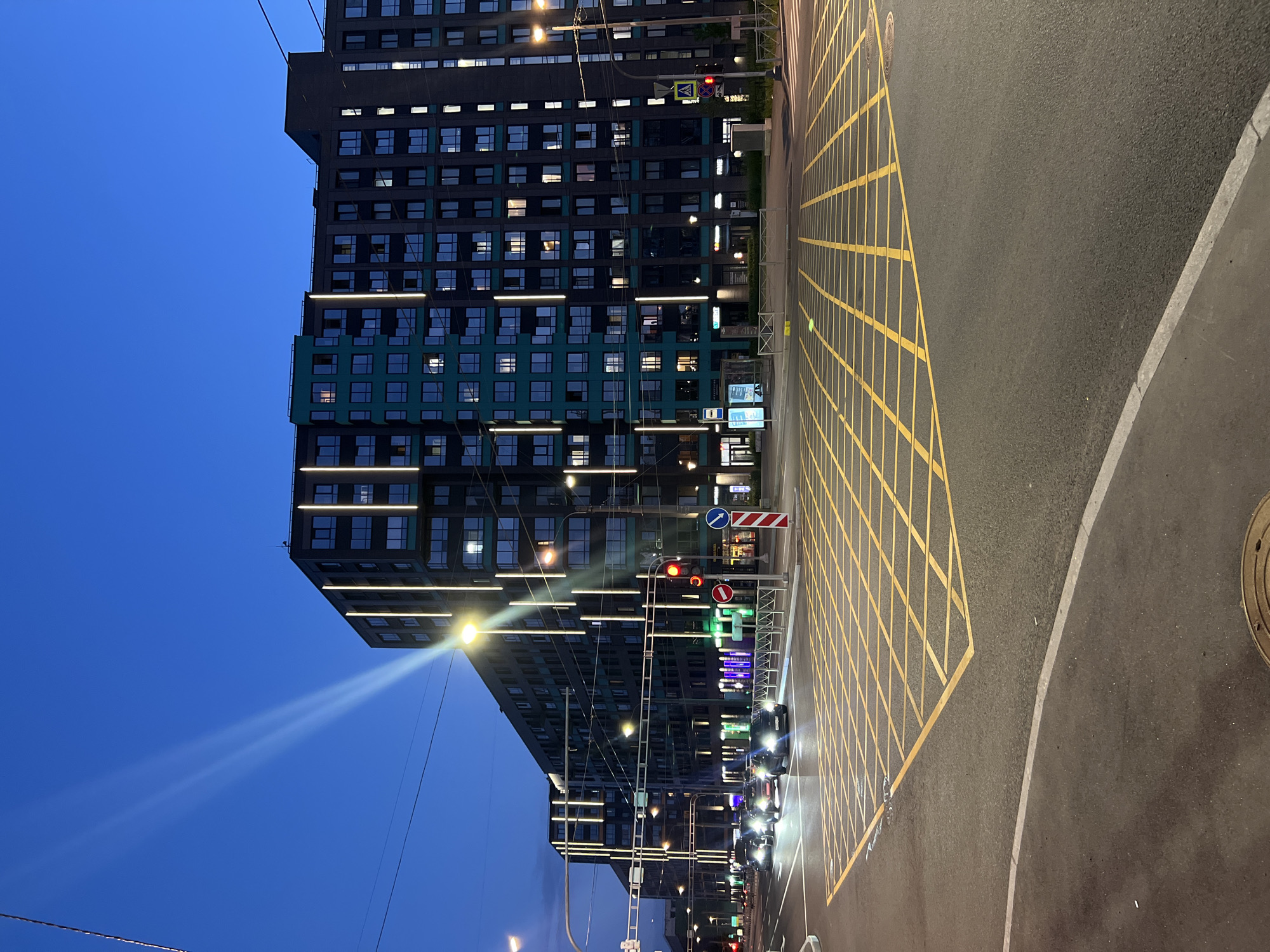This nocturnal urban street scene, situated in Saint Petersburg, Russia, depicts an intersection bordered by contemporary multi-story buildings under a clear, dark blue sky. The dominant structures are modern residential or mixed-use complexes, characterized by dark gray facades, interspersed teal-colored panels, and distinctive vertical white light strips illuminating their exterior. Numerous windows within these buildings show interior lighting.
The asphalt road in the foreground features a prominent yellow box junction grid, along with white solid and broken lane markings. Four dark-colored sedan-type cars are positioned in the left lanes, facing away from the viewer, with their headlights illuminated. Their state suggests they are either stopped or moving slowly towards the intersection.
A red circular traffic light is clearly visible on a pole on the right side of the intersection, indicating 'stop' for vehicular traffic. Attached to the same pole is a white triangular pedestrian crossing sign. Overhead, multiple utility wires traverse the frame. Several tall streetlights emit bright yellow-white light, illuminating the road and building facades. Additional street furniture includes a bus stop shelter with advertising panels on the left, low metal pedestrian barriers along the sidewalk, and a red-and-white striped regulatory sign (likely 'no stopping' or 'no parking') near the traffic light pole. No individuals are discernible within the image.