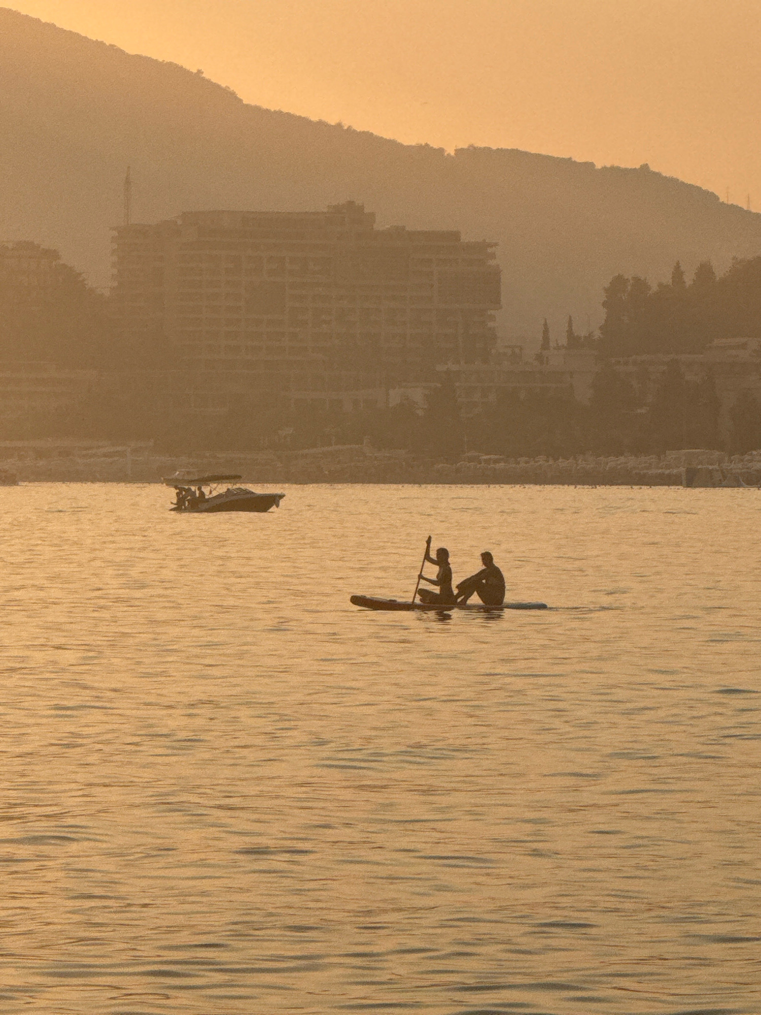 The image depicts a serene scene on the water in Rafailovići, Montenegro, during a sunset or sunrise, characterized by a warm, golden hue that bathes the entire landscape. In the foreground, two figures are silhouetted on a paddleboard, one seated and holding a paddle, while the other is seated behind them. Their forms are indistinct against the vibrant water. Further in the background, a motorboat with several occupants is visible, cutting through the gentle ripples of the sea. The middle ground reveals a shoreline dotted with buildings, including a prominent, multi-story structure that appears to be a hotel or resort, its architecture suggesting a developed coastal area. Beyond the structures, the silhouette of a mountain range rises, softened by atmospheric haze and the low-angled light. The overall impression is one of tranquility and natural beauty, with human activity present but harmoniously integrated into the environment.