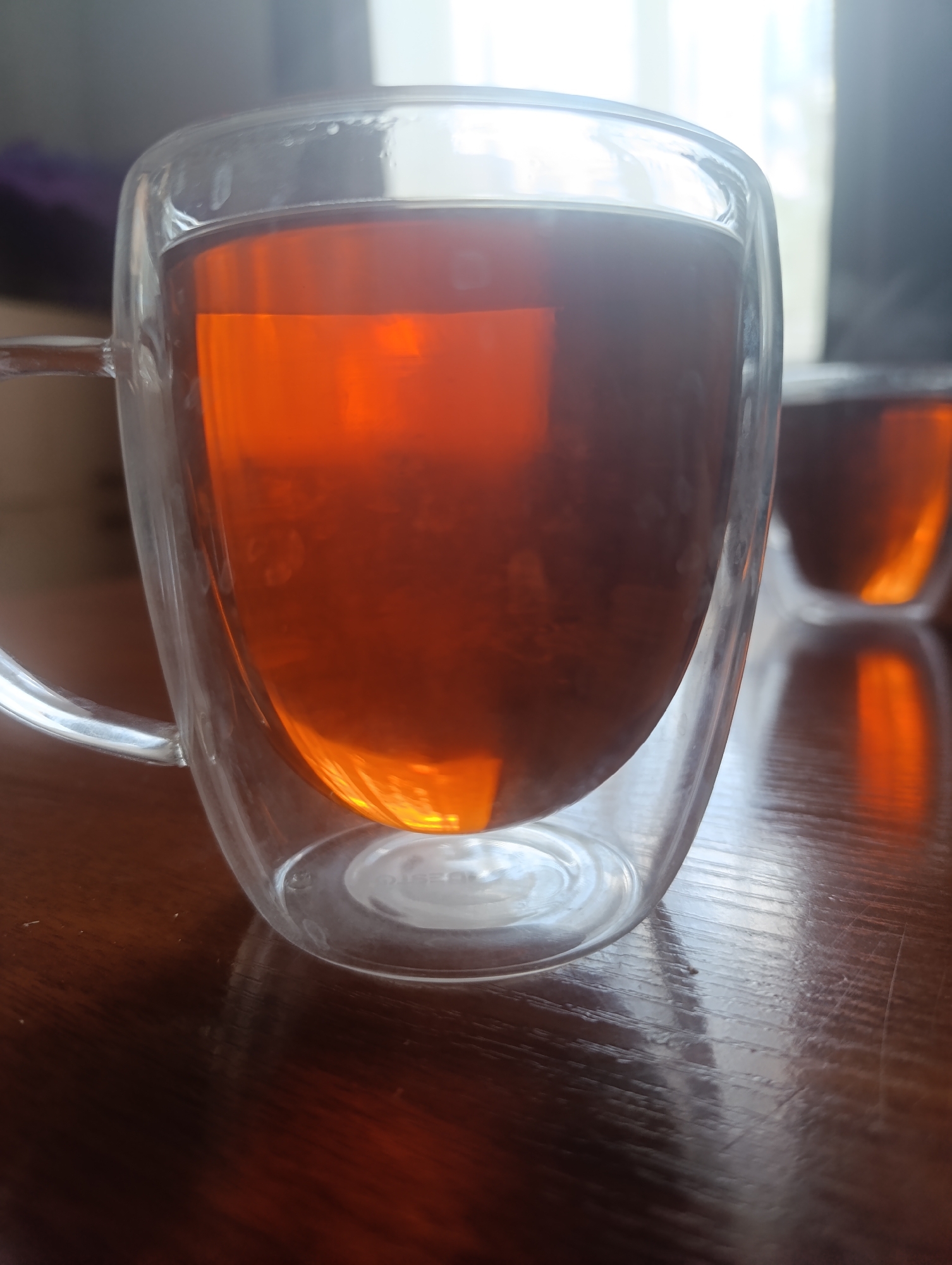 A close-up, eye-level shot shows a double-walled glass mug filled with amber-colored liquid, likely tea, on a dark wooden table. The mug has a clear glass handle. The liquid inside is translucent and reflects light, creating bright orange and yellow hues. The mug is positioned slightly off-center to the left of the frame. In the background, the scene is out of focus, suggesting a bright window or light source. A second, similar mug is partially visible to the right, also filled with the same liquid. The overall impression is one of warmth and relaxation. The location is Unknown, Unknown.
