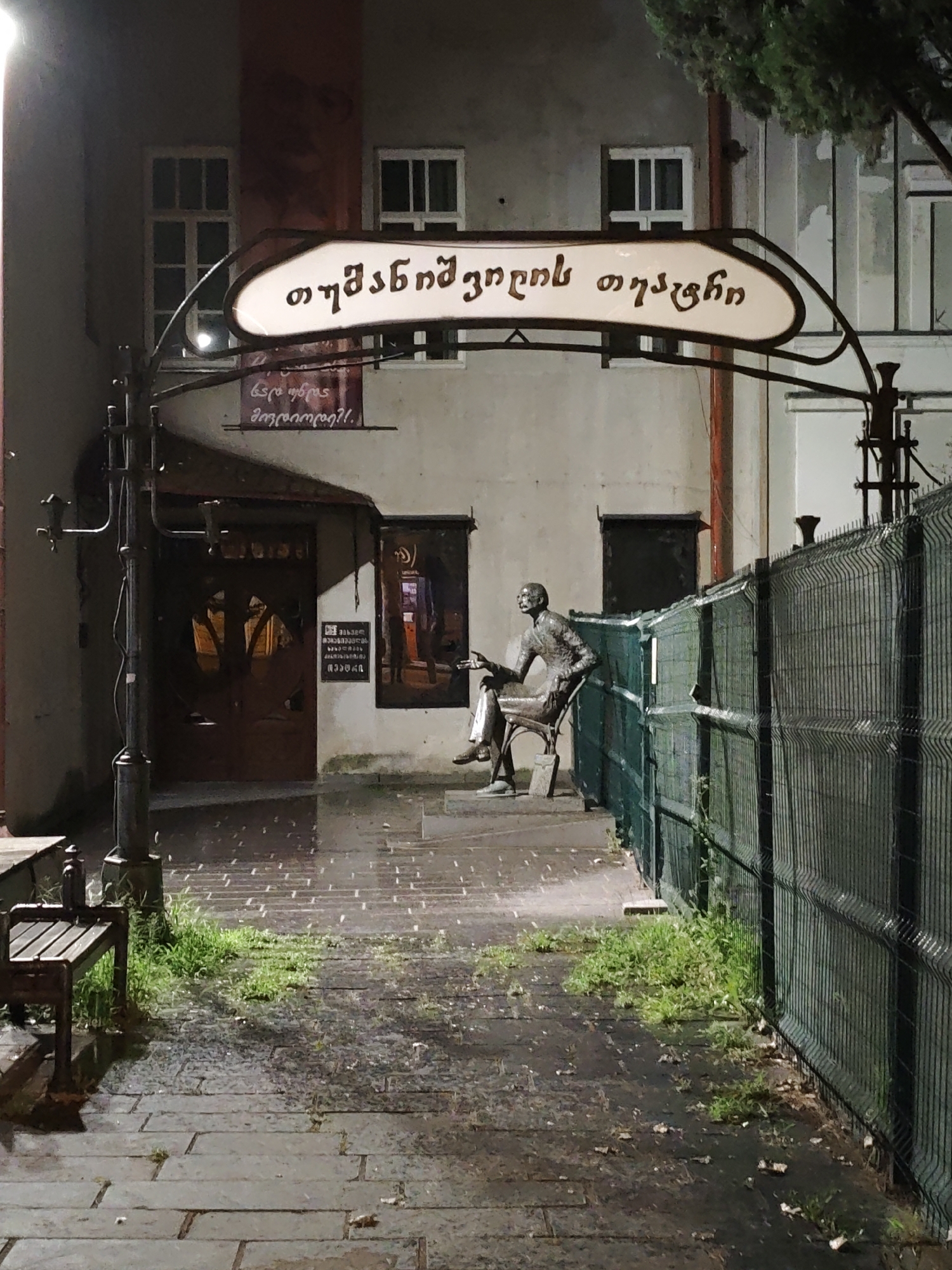The image depicts the exterior of a building at night in Tbilisi, Georgia. A curved metal sign with Georgian script is visible above an entryway. To the right of the entryway, a bronze statue of a seated man is positioned behind a green chain-link fence. The ground in front of the building is paved with large, flat stones, and grass grows in patches. A wooden bench is situated to the left of the entryway. The lighting suggests it is nighttime, and the ground appears damp, indicating recent rain. The overall atmosphere is somber and quiet.
