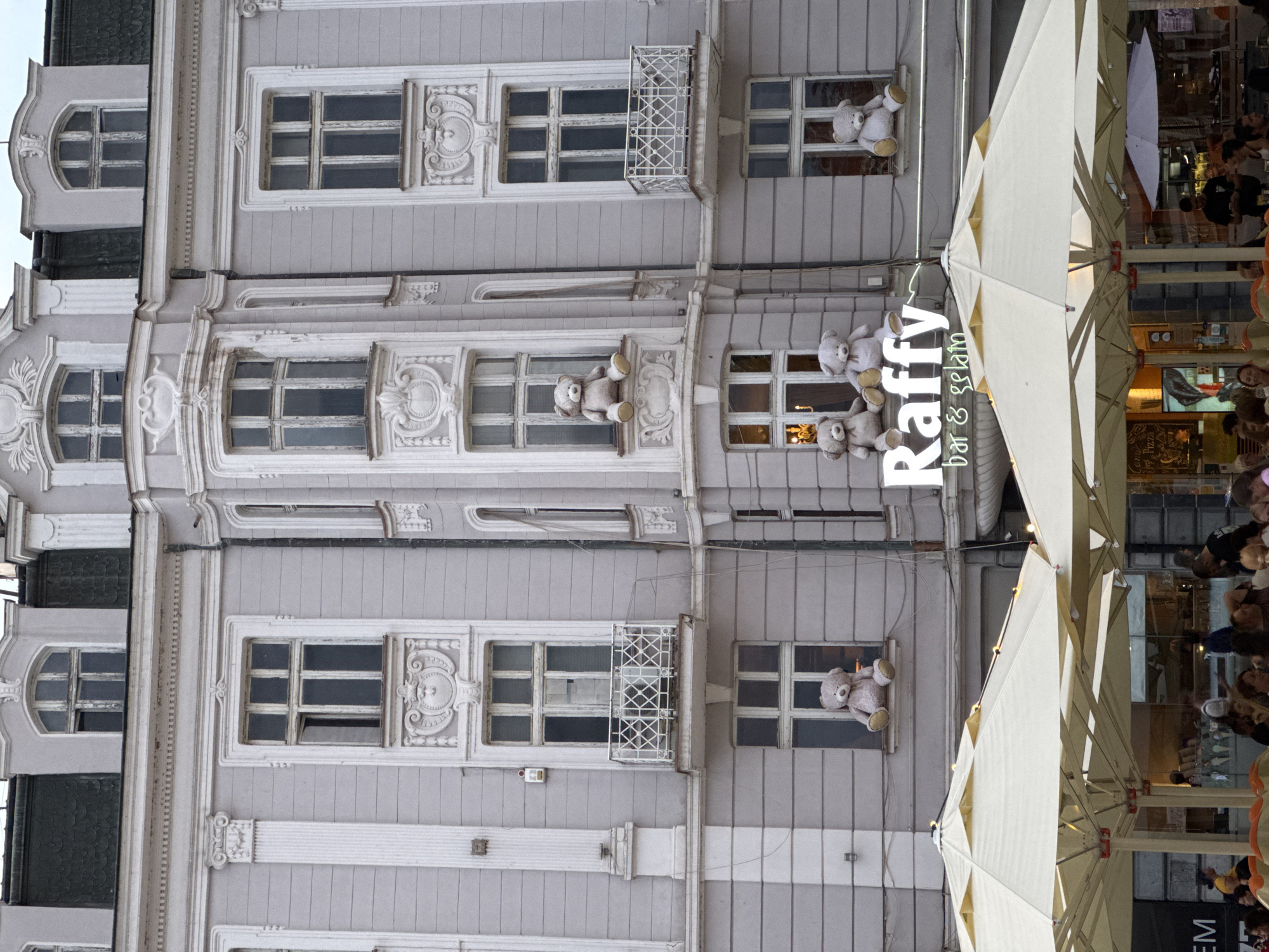 This is a photograph of the facade of a light gray building in Plovdiv, Bulgaria, with several teddy bears placed in the windows. The building features ornate architectural details, including columns, decorative moldings, and balconies. From left to right, a large light brown teddy bear is seated on the windowsill of a window on the fourth floor, appearing to look out. Below it, on the third floor, two more teddy bears, one light brown and one off-white, are seated on the windowsill. To the right of these, on the fourth floor, another large light brown teddy bear is seated on the windowsill. Below this, on the second floor, a balcony with decorative white railings is visible. The building has multiple stories with numerous windows, many of which are large and multi-paned. A sign for "Raffy bar & gelati" is visible on the building, below the teddy bears. In the foreground, the tops of cream-colored outdoor umbrellas and tables are visible, suggesting a cafe or restaurant setting. People are seated at some of the tables in the background, partially obscured by the umbrellas. The lighting suggests it is daytime, with overcast conditions.