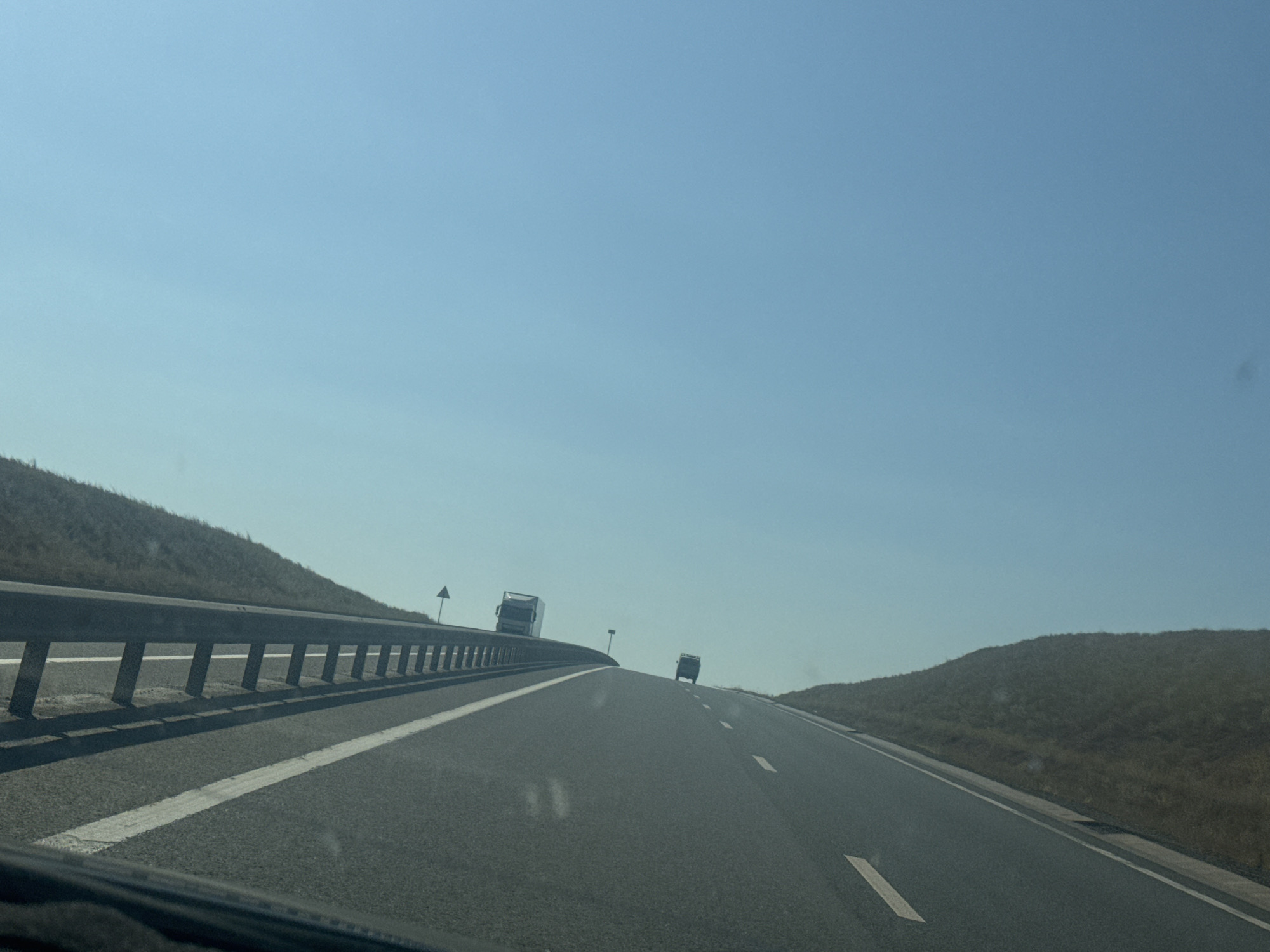 The image captures a clear, sunny day on a highway. The perspective is from inside a vehicle, looking forward. A guardrail lines the left side of the road, separating it from a grassy embankment. The highway itself has two lanes, marked by white lines. In the distance, two trucks are visible, driving in the same direction as the viewer. The sky is a pale blue, with no clouds. The terrain appears to be rolling hills covered in dry grass. There is no indication of this being in the city of Syrt, Russia; it looks like a rural highway.
