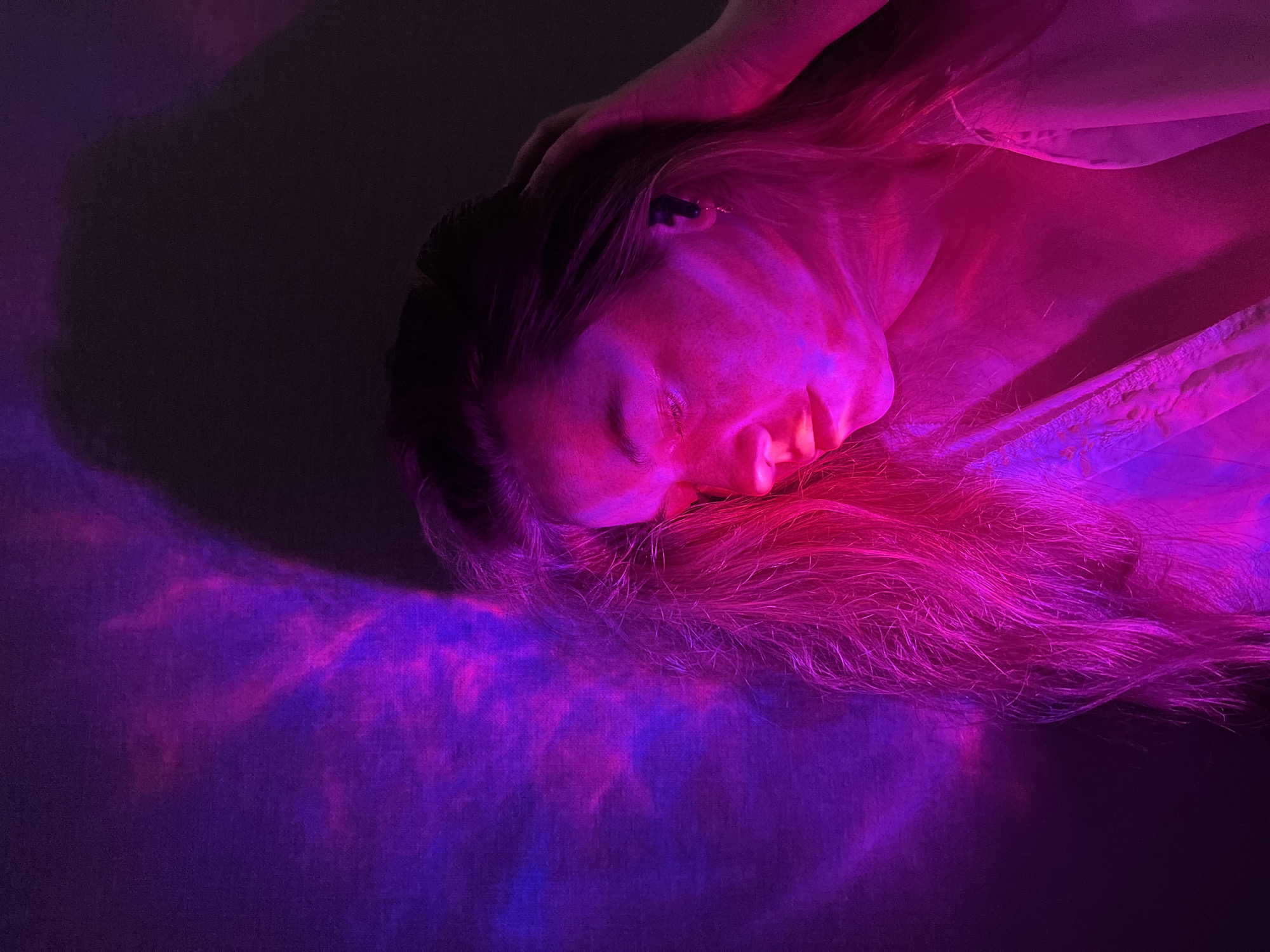 A person with long blonde hair is lying down, illuminated by pink and purple light. The person's face is turned to the side, and their eyes are closed. Their hand is resting on their head. The background is a dark surface with purple light patterns. There is no indication of the city of Chaikovsky, Russia.