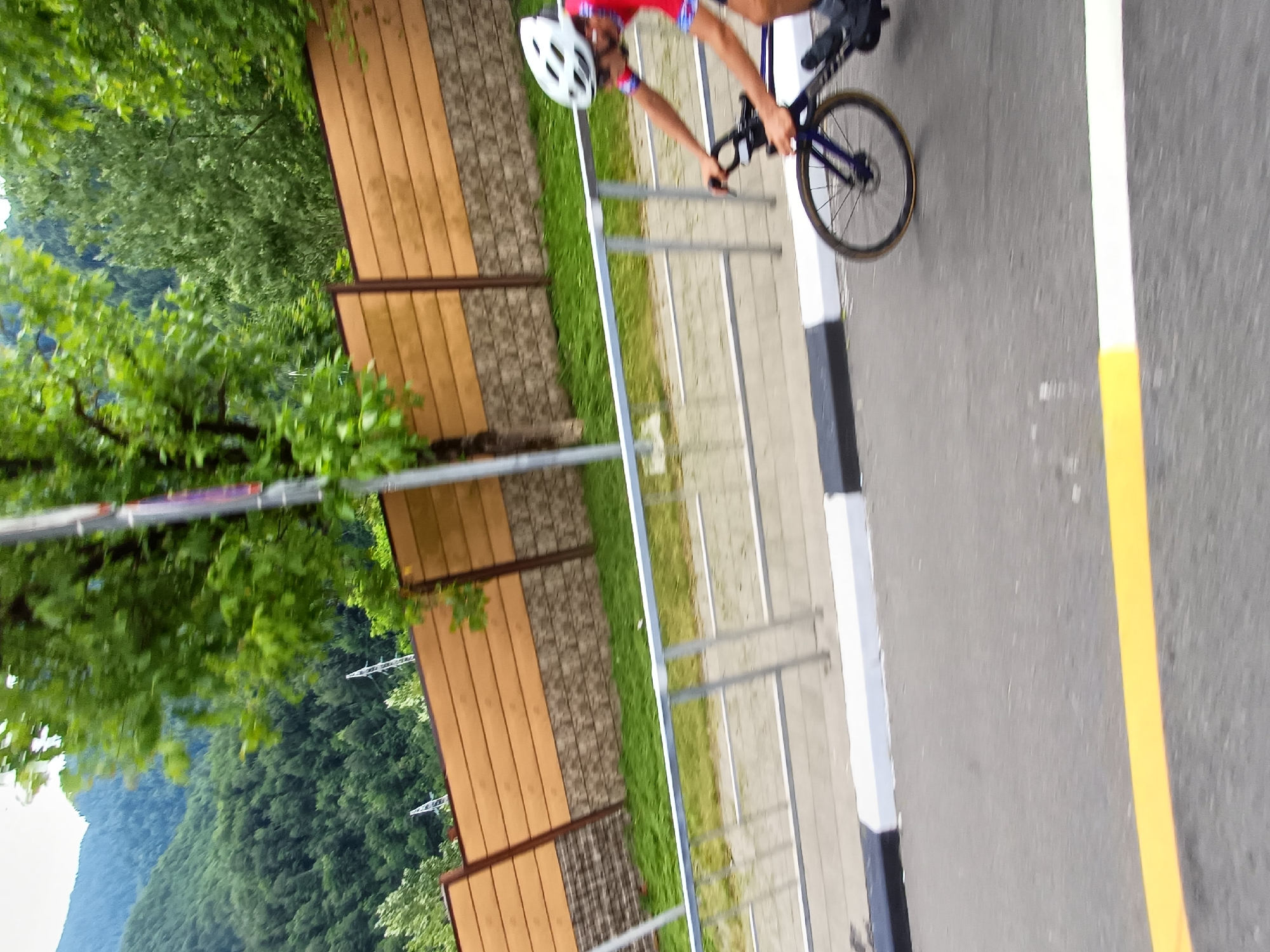 A cyclist wearing a white helmet and a red and blue cycling outfit is captured in motion, riding a dark blue road bike. The cyclist is positioned in the mid-right of the frame. The background features a wooden fence and lush green trees. In the distance, a mountain range covered in dense green foliage is visible under a cloudy sky. The scene appears to be on a road with a black and white striped curb and a yellow line painted on the asphalt. The image quality suggests it was taken with a mobile device, possibly during a sporting event or recreational activity in Krasnaya Polyana, Russia, known for its mountainous terrain and outdoor activities.