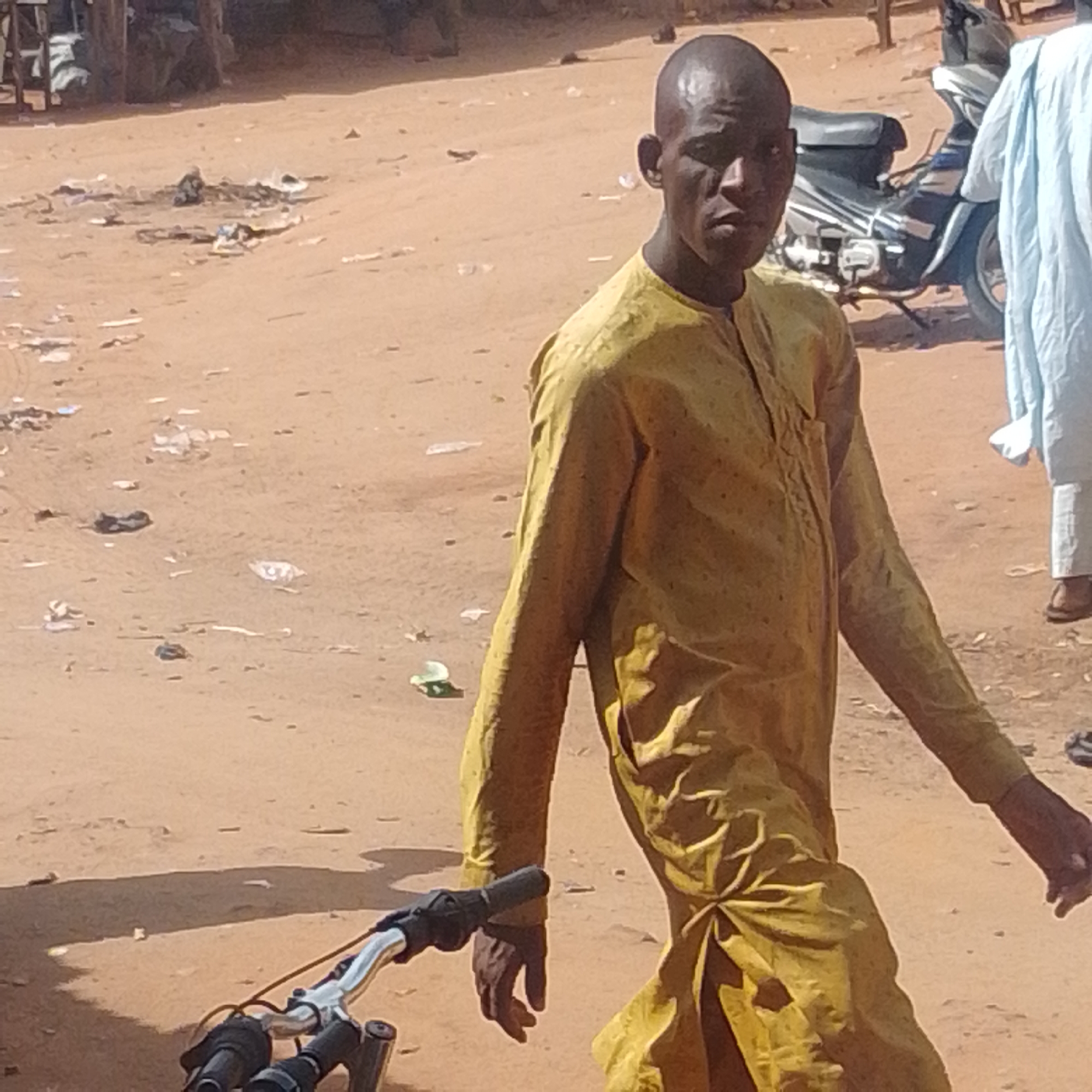 In Gombe, Nigeria, a man in a yellow robe walks across a dirt path, his bicycle handlebars visible in the foreground. He appears to be walking towards the right side of the frame, his gaze directed forward with a neutral expression. The ground is dusty and littered with debris, suggesting an outdoor, possibly rural or undeveloped, setting. Behind him, a motorcycle is parked, and another person, partially visible and wearing a light blue garment, stands in the background. The lighting suggests it is daytime, with the sun casting shadows, indicating a sunny day. The overall scene depicts a moment of everyday movement in a dusty, possibly arid environment.