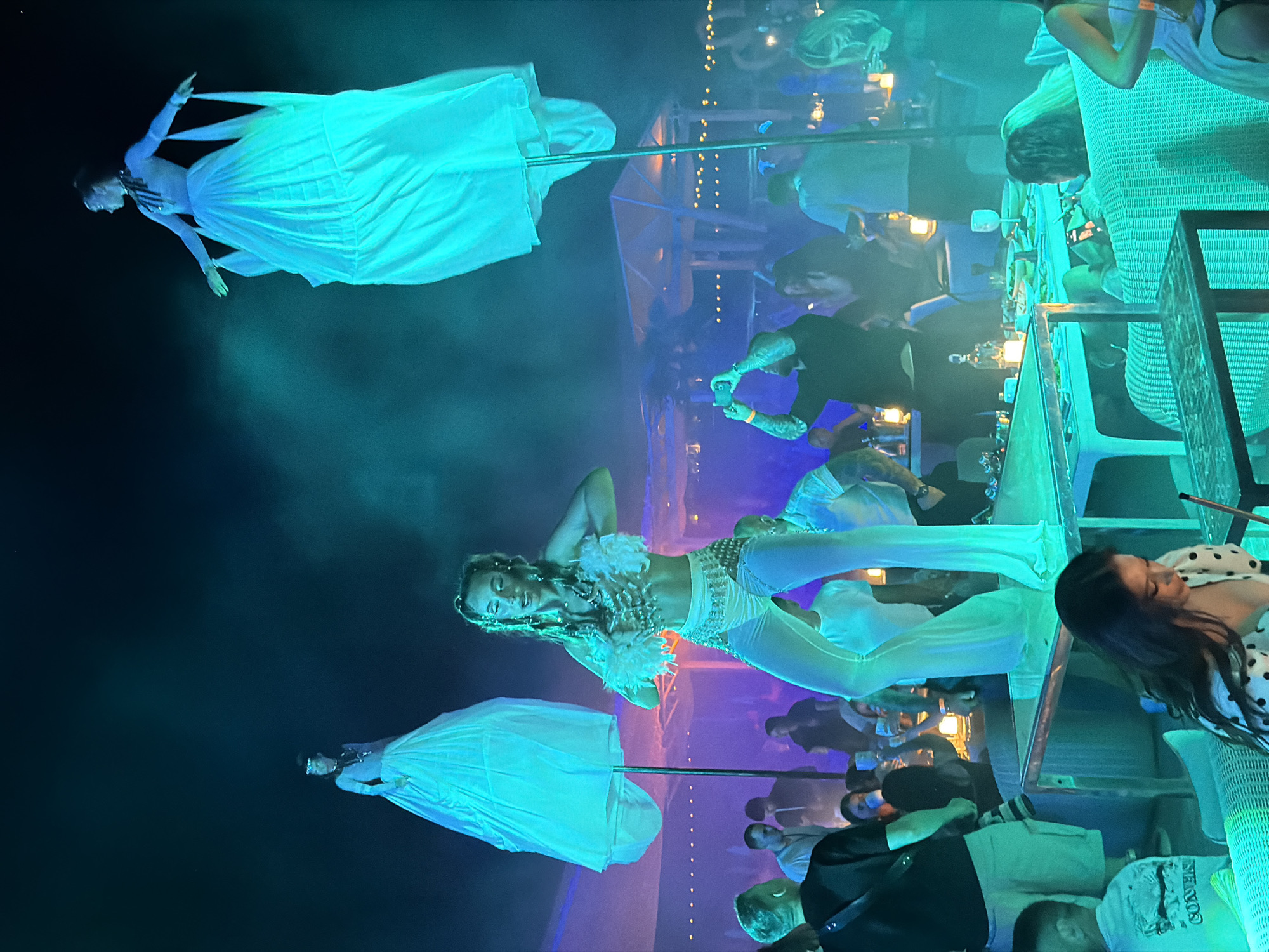 The image depicts a vibrant nighttime outdoor event, likely a performance or festival, illuminated by teal and purple lighting, set against a dark sky. Three performers are prominently featured, each standing on elevated platforms. The performer in the foreground, center, is a woman dressed in white, sequined attire, including a cropped top with feather accents and bell-bottom pants. She is captured in a dynamic pose, with one leg extended and arms raised, suggesting a dance or acrobatic movement. To the left and right, two other performers are elevated on tall, thin poles, wearing voluminous, floor-length white dresses that billow around them, giving them an ethereal appearance. They are also posed, with arms outstretched.
The background reveals a lively crowd of people seated at tables, interspersed with standing individuals, suggesting a social gathering, perhaps at a restaurant or lounge. The environment appears to be a beach club or an open-air venue, with glimpses of white lounge furniture and tables set with glassware and bottles. The overall atmosphere is festive and energetic, with a sense of theatricality due to the performers' elaborate costumes and staging. The location is Odesa, Ukraine.
