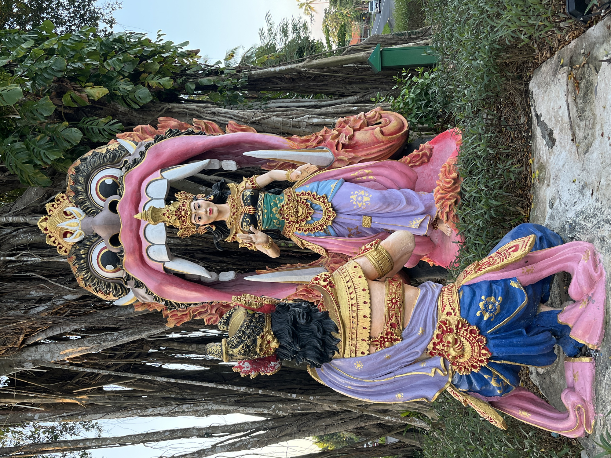 A photograph captures a vibrant and ornate statue installation, likely a religious or cultural monument, situated outdoors in Kuta Selatan, Indonesia. The scene is dominated by two large, brightly colored statues, one positioned in the foreground and the other behind it.
The statue in the foreground is kneeling and viewed from the back. It depicts a male figure adorned in elaborate traditional attire. He wears a golden crown with red embellishments, a golden chest plate with intricate patterns, and a blue lower garment that drapes around his legs. A purple sash is draped over his left shoulder and across his torso, fastened with a golden belt featuring a circular, sun-like emblem. His knees are bent, and his hands are not fully visible.
Behind the kneeling figure is a much larger, more imposing statue. This statue features a monstrous, gaping mouth with large white teeth and a bright pink interior. Within the mouth, a female figure stands regally. She is also dressed in ornate traditional clothing, including a golden crown, a green and gold bodice, and a flowing skirt in shades of pink and purple. Her hands are raised, with the right hand in a gesture of blessing or protection. The entire structure is set against a backdrop of a large, ancient tree with numerous hanging aerial roots, characteristic of banyan trees common in tropical regions.
The surroundings suggest a public space or a roadside attraction. Greenery, including manicured bushes and some fallen leaves, surrounds the statues. A paved road with a white line marking the lanes is visible in the background to the right, with a car driving away from the camera. A small green post or bin stands near the edge of the road, partially obscured by foliage. The sky above is clear and blue, indicating daytime. The overall impression is one of cultural richness and artistic expression, with the statues serving as a focal point in the tropical landscape of Kuta Selatan.