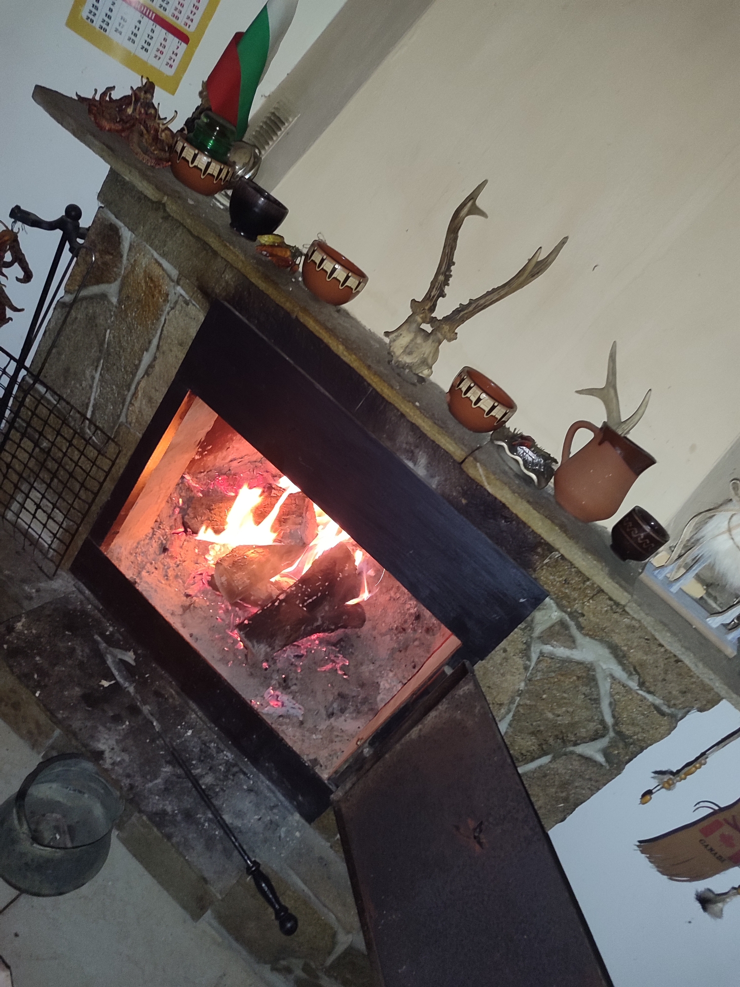 The image depicts a stone fireplace with a fire burning inside. On the mantel above the fireplace are decorative items, including antlers, pottery, and a Bulgarian flag. A calendar hangs on the wall to the left of the flag. A metal grate and a black pot sit to the left of the fireplace opening, along with a poker. The photograph appears to be taken indoors, with a warm, inviting atmosphere created by the fire. While the location cannot be definitively stated, the presence of the Bulgarian flag suggests that it could be in Bulgaria, perhaps in Varshilo.
