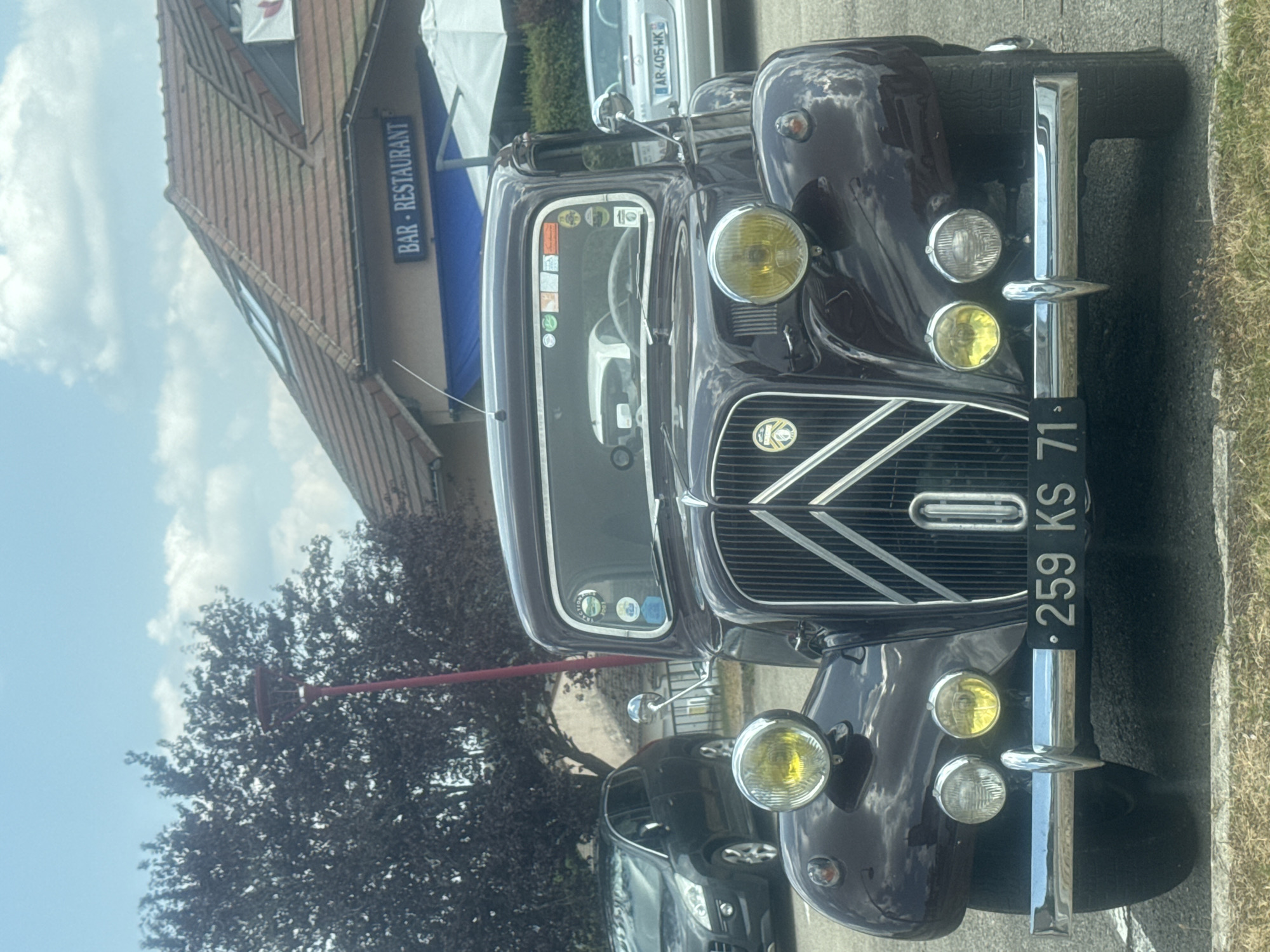 The image shows a vintage dark purple Citroën Traction Avant parked in front of a building with a "BAR - RESTAURANT" sign. A dark grey Peugeot hatchback is parked to the left of the Citroën, and a silver convertible with license plate "AR 405 WK" is visible to the right. The scene appears to be outdoors on a sunny day with scattered clouds. The Citroën has a license plate reading "259 KS 71". The building has a tiled roof and a blue awning. The foreground shows grass and pavement. The image is cropped to focus on the front of the vintage car.