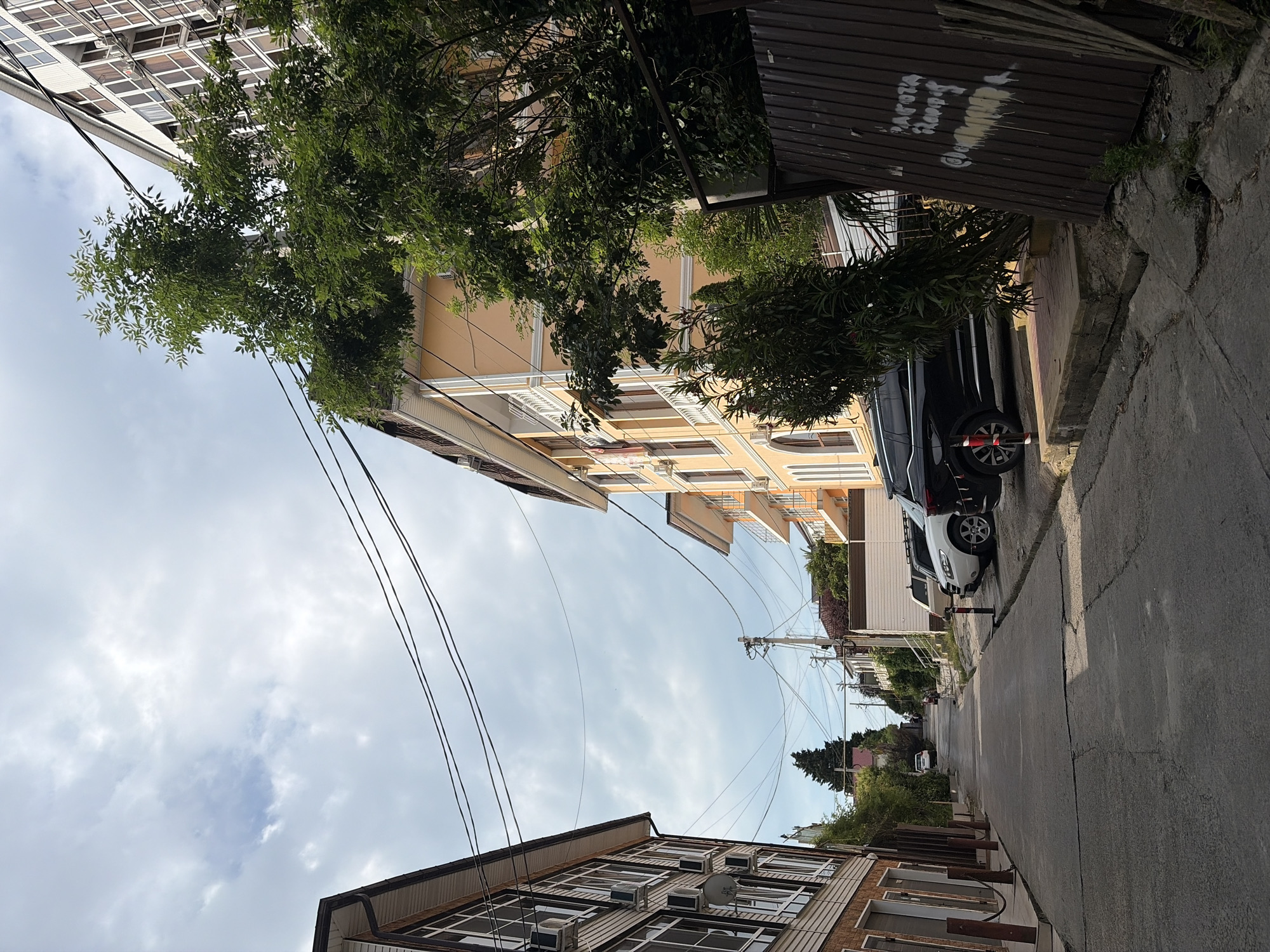 The image shows a narrow street in Sochi, Russia, with multi-story buildings on either side. The street is paved and appears to be a residential area. Several cars are parked along the side of the street. There are trees and greenery visible, as well as power lines overhead. The sky is cloudy.