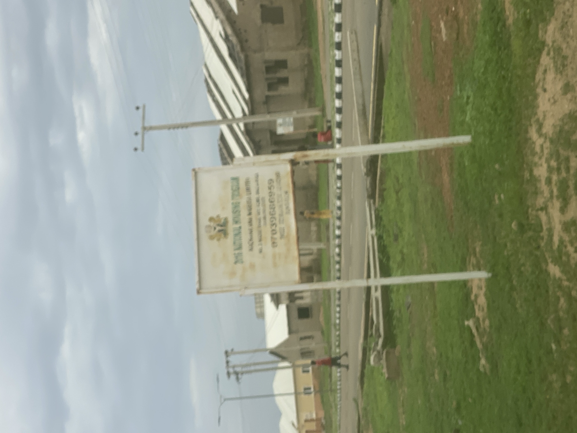 The image captures a scene in Dumnga, Nigeria, featuring a signpost for "2016 NATIONAL HOUSING TRUST." The sign, white with some text and an emblem at the top, is supported by two white poles. Behind the signpost, several houses with corrugated iron roofs are visible, some appearing unfinished or in a state of disrepair. A paved road with a black and white striped curb runs in front of the houses and past the sign. In the foreground, there is a patch of green grass and some dirt. A person in a red shirt is walking across the road, and another person in a yellow garment is standing near the houses. Utility poles and streetlights are also present in the background, suggesting a residential or developing area. The sky is overcast with clouds.
