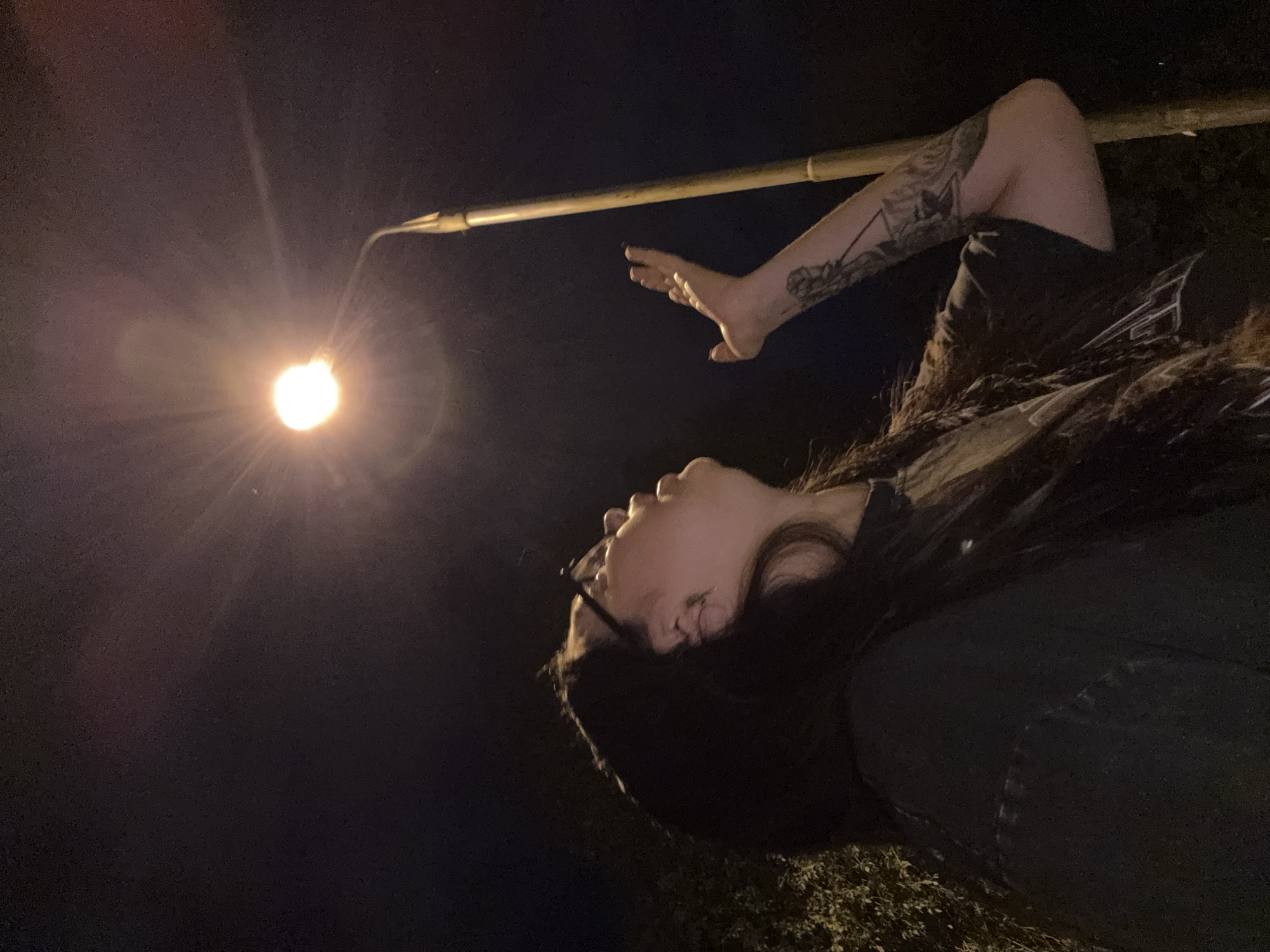 A person with long, dark hair and glasses is reaching their left hand up towards a streetlamp at night. The person is wearing a dark t-shirt. A tattoo is visible on their left forearm. The background consists of dark foliage and a tall, light-colored pole supporting the streetlamp. The image is captured at street level.