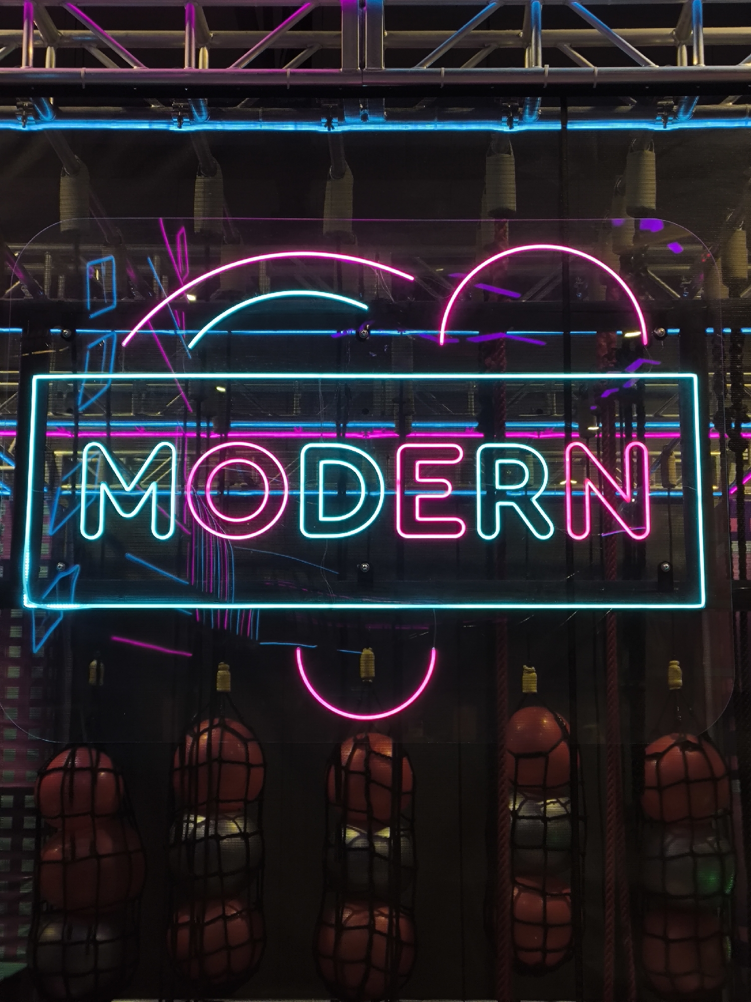 The image shows a neon sign spelling out "MODERN" in a combination of cyan and pink neon tubes. The sign is set against a dark background with various metal structures and further neon lighting in blue and pink. Below the sign, several netted bags filled with red and grey spherical objects are hanging. The overall aesthetic suggests an entertainment venue or a modern art installation. The provided location of Орловка, Russia, is not visually confirmed by any elements within the image itself.