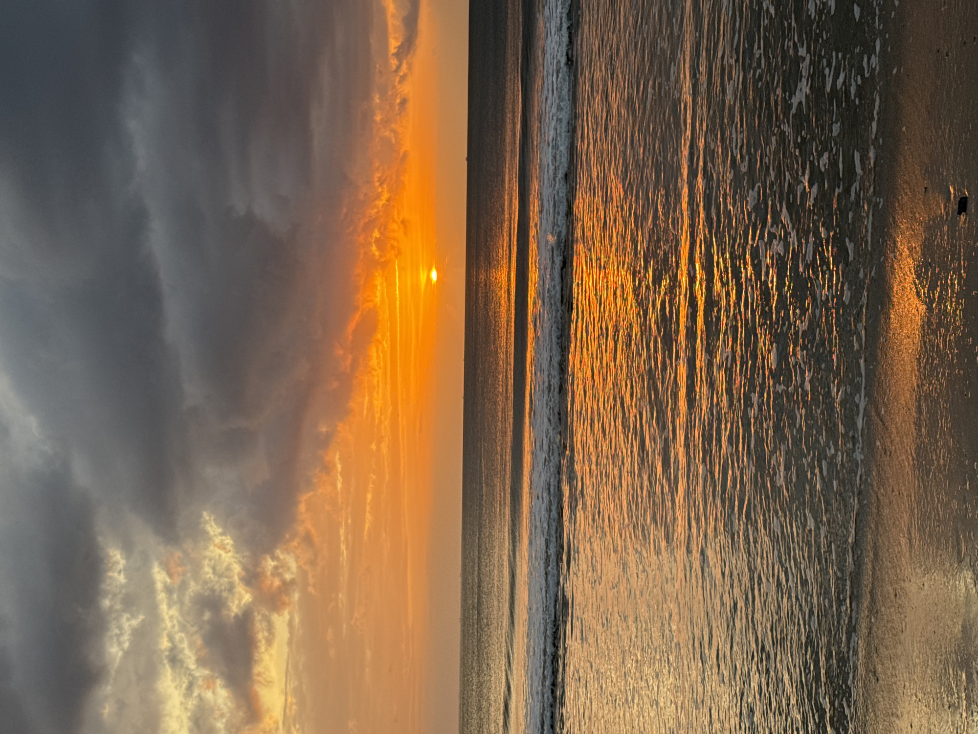 This is a landscape photograph of a sunset over the ocean, likely taken from a beach. The sky is filled with vibrant orange and yellow hues from the setting sun, contrasted with dark, dramatic clouds. The sun itself is a small, bright disk visible on the horizon. The ocean reflects the colors of the sky, with gentle waves breaking on the shore. The wet sand in the foreground also reflects the warm light. Based on the presence of typical Indonesian fishing boats visible in the distance on the ocean, and the style of the sunset, the location is identified as Kuta Selatan, Indonesia. There are no people or animals visible in the frame. The scene depicts a natural environment at dusk.