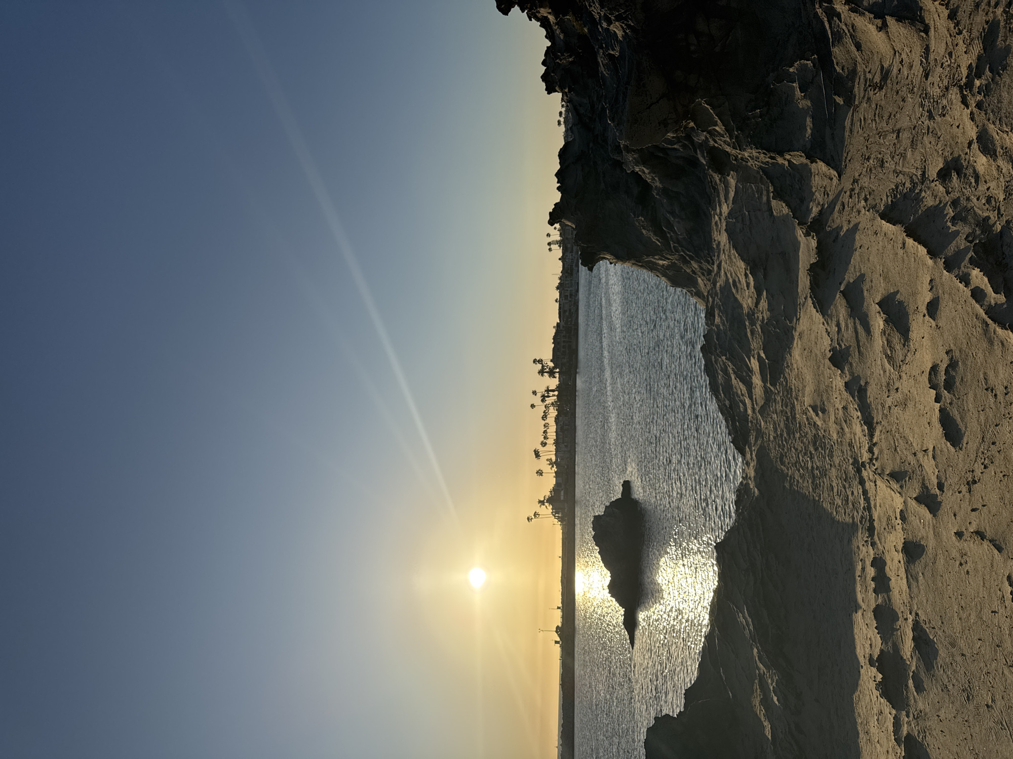 The image depicts a beach scene in Newport Beach, California, USA. The sun is low on the horizon, emitting bright rays across the sky and reflecting on the water. The coastline is lined with palm trees. A rocky outcrop, possibly a small island or peninsula, is visible in the water, reflecting the sunlight. The foreground shows a rocky cliff with sandy texture. The sky is a gradient of blue to yellow, indicating either sunrise or sunset.