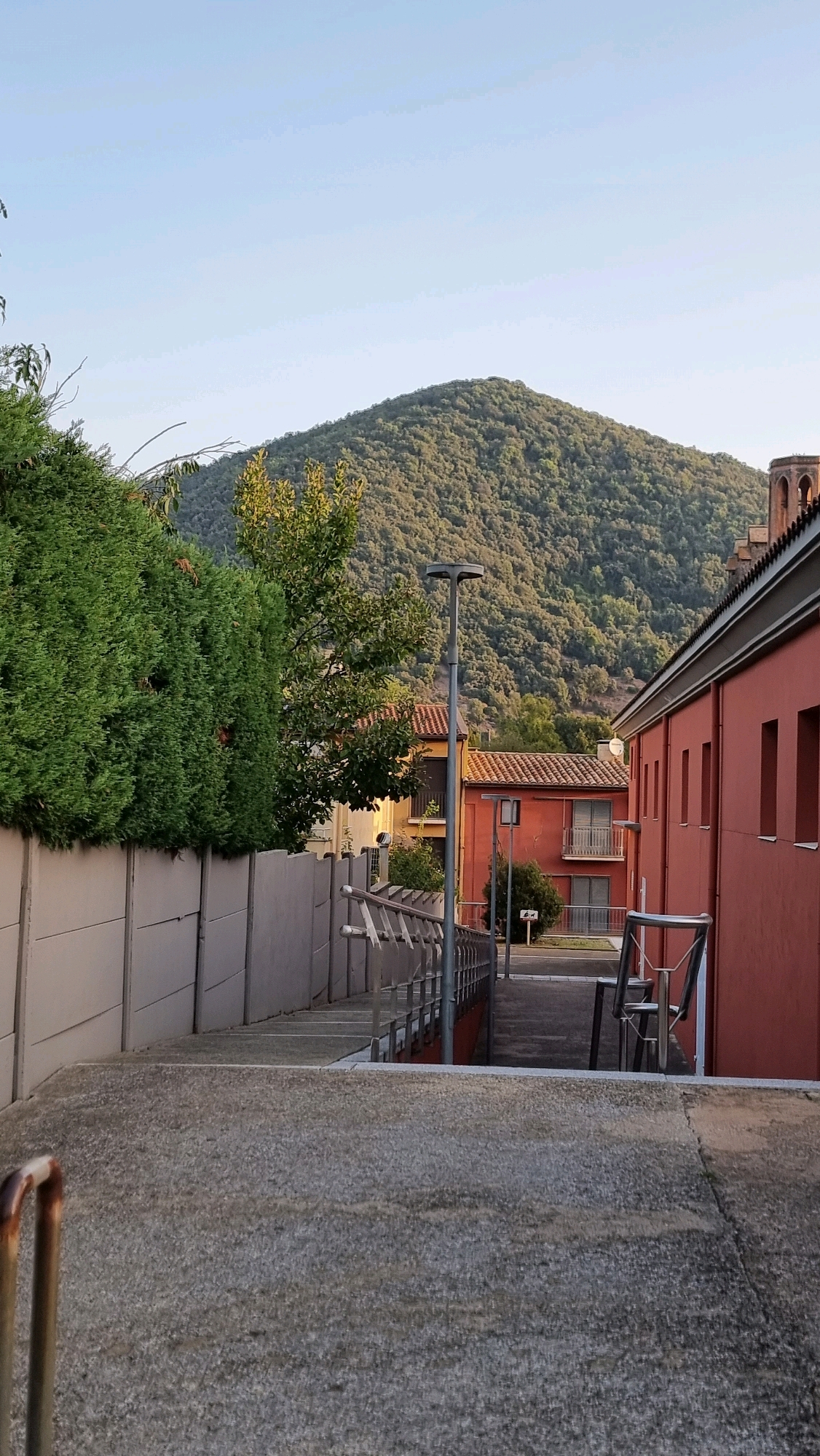 The image shows an outdoor view with a prominent green hillside in the background, covered in dense foliage. In the middle ground, there are several buildings. One building on the right is painted a vibrant reddish-orange and has a series of vertical rectangular windows. Adjacent to it, a smaller building with an orange tiled roof features balconies. To the left, a thick, well-maintained green hedge forms a barrier along a light-colored concrete wall. A modern street lamp with a circular top stands between the hedge and the buildings. In the foreground, a concrete staircase with a metal handrail leads downwards. The sky is clear and pale blue, indicating daylight. The scene appears to be a residential or communal area, possibly a quiet street or pathway leading to buildings. The image does not contain any people or animals. The location is identified as Sant Joan Les Fonts, Spain.