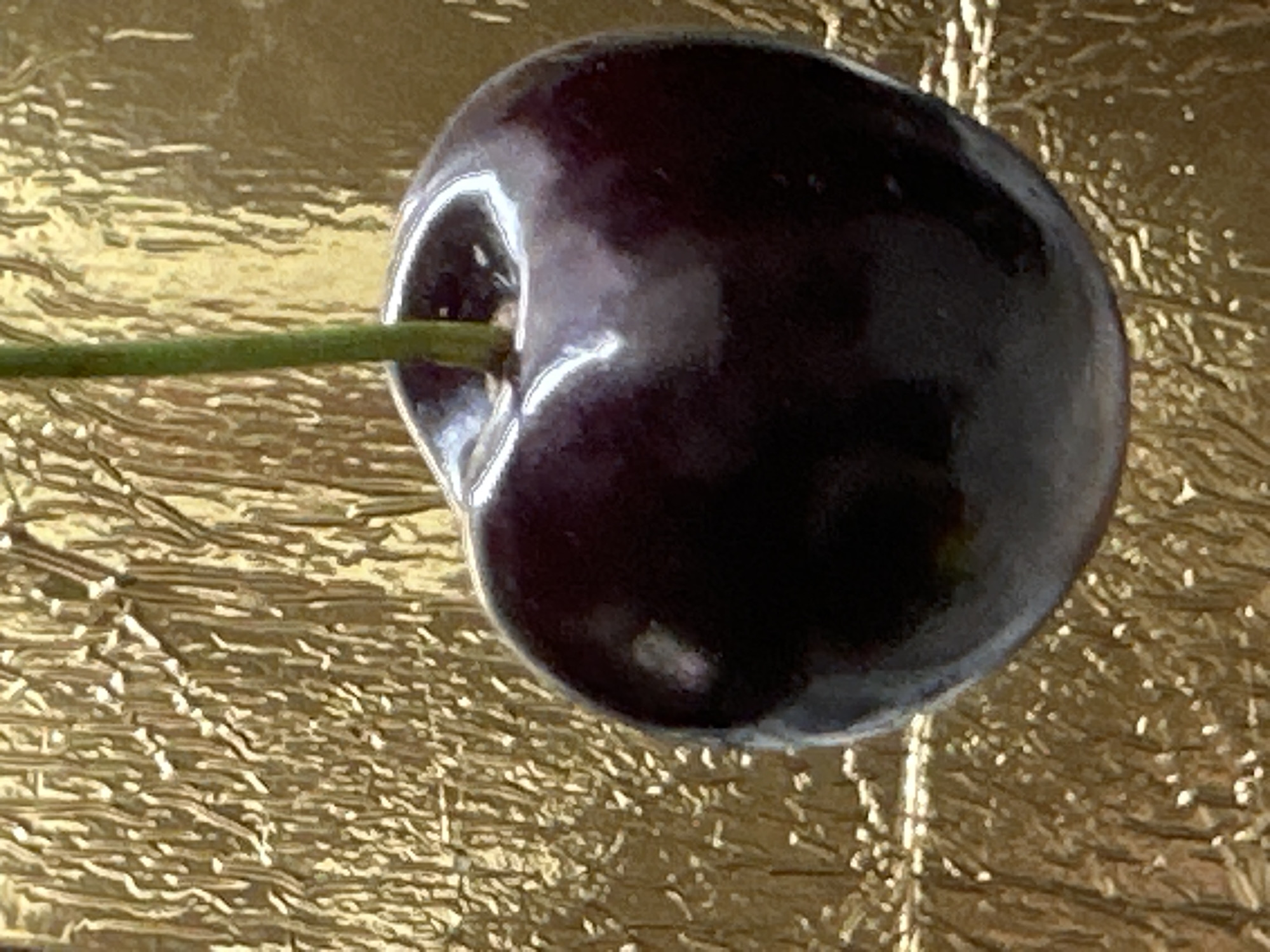 This is a close-up, portrait-oriented photograph of a single, dark red cherry with a thin green stem against a textured, metallic gold background. The cherry is glossy and reflects light, showing highlights on its surface. The stem is positioned vertically, emerging from the top center of the cherry and extending upwards out of the frame. The gold background is crinkled and uneven, creating a pattern of light and shadow. The image does not depict any specific location within the city of Luhansk, Ukraine. There are no people, actions, or interactions visible in the photograph.