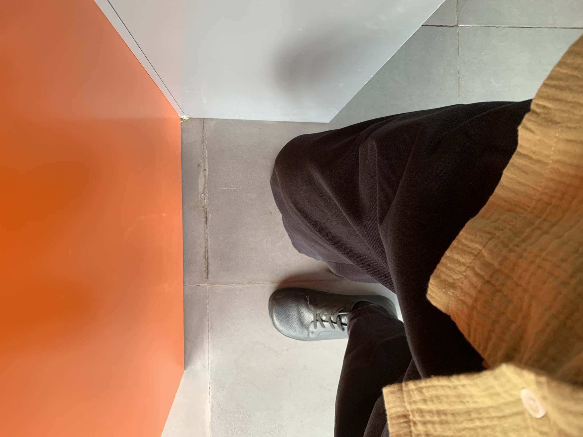 The image is a close-up, overhead shot of a person's legs and feet, viewed from the perspective of someone standing or sitting. The person is wearing black pants and a black shoe. A portion of a tan-colored shirt is visible on the right side of the frame. The scene appears to be indoors, with a tiled floor. To the left, there is a vertical orange surface and a white surface above it, suggesting a wall or partition. The lighting is consistent and suggests an indoor setting with artificial light or diffused natural light, rather than a specific time of day. There are no discernible emotions, weather conditions, or visible text in the image. The location context provided is Megion, Russia, but this information cannot be visually confirmed from the image itself.