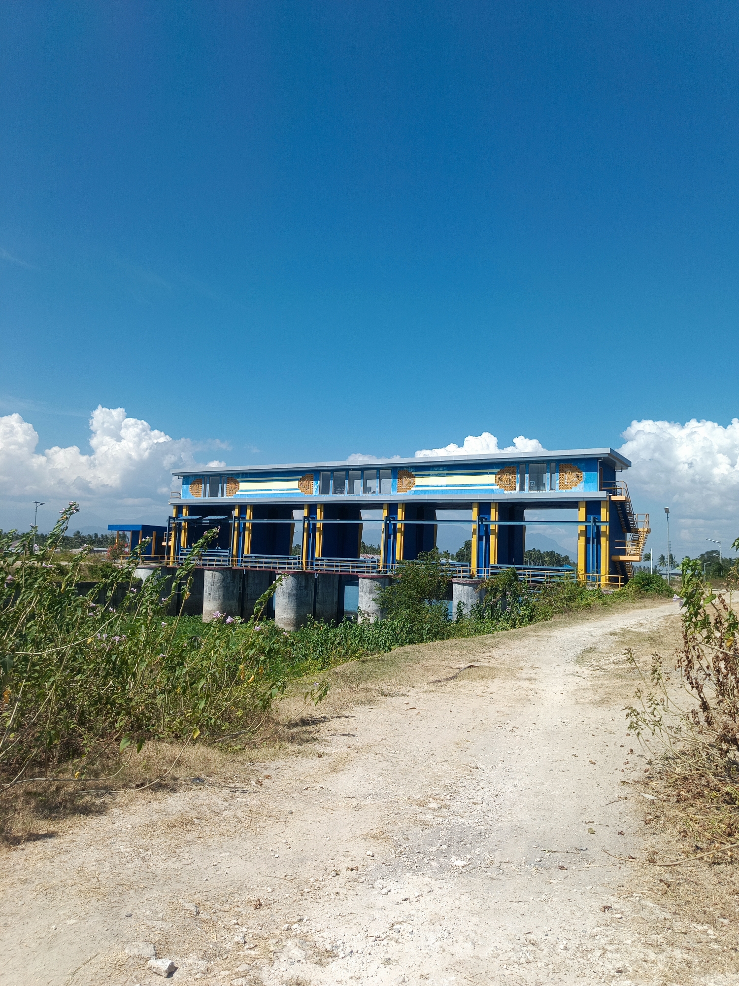 The image depicts a concrete dam structure with a bright blue and yellow superstructure, situated in Ilotidea, Indonesia. The dam is flanked by large, cylindrical concrete pillars supporting the main structure, which features several large openings. The superstructure is a long, rectangular building with a flat roof, adorned with decorative elements in yellow and blue. Staircases with yellow railings provide access to the upper levels.
In the foreground, a dirt path, appearing dry and dusty, leads towards the dam. The path is bordered by vegetation, with dry grasses and some green plants with small purple flowers visible. To the left of the path, dense green foliage and bushes are prominent, partially obscuring the base of the dam.
The sky above is a vibrant, clear blue, with a few scattered white cumulus clouds, some of which are quite large and puffy. In the distance, beyond the dam and to the left, a hazy landscape with distant hills or mountains can be seen, along with some trees and buildings, suggesting a populated area. The overall impression is one of a sunny day in a rural or semi-rural setting. There are no people visible in the image. The scene is static and depicts a fixed structure within its environment.