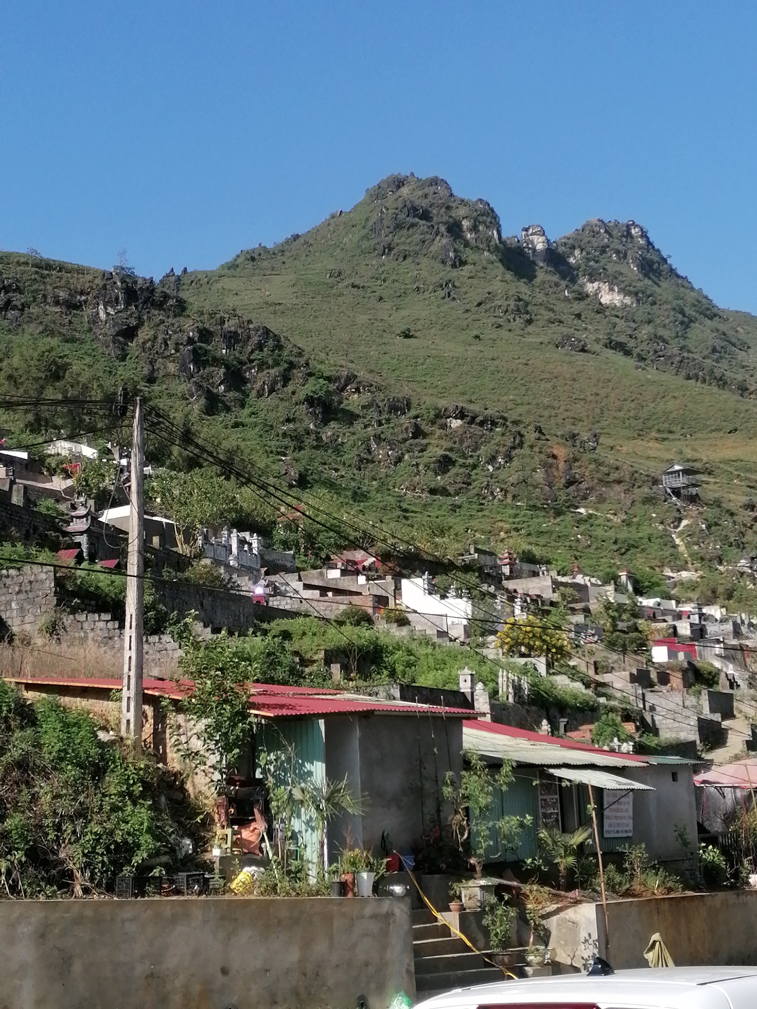 The image shows a hillside settlement in the Sa Pa Ward, Vietnam. A large, green mountain dominates the background, receding into a clear blue sky. The hillside is densely populated with small buildings, primarily simple dwellings with red and green roofs. Stone retaining walls define the different levels of the hillside. A concrete utility pole, from which electrical wires run, is prominent on the left side of the frame. In the foreground, a low concrete wall runs horizontally across the frame, in front of a house with a green roof. There are plants and other objects situated on the stairs in front of it. The top of a white car peeks into the frame at the bottom right. Overall, the scene portrays a rural, mountainous settlement with a mix of natural and built environments.