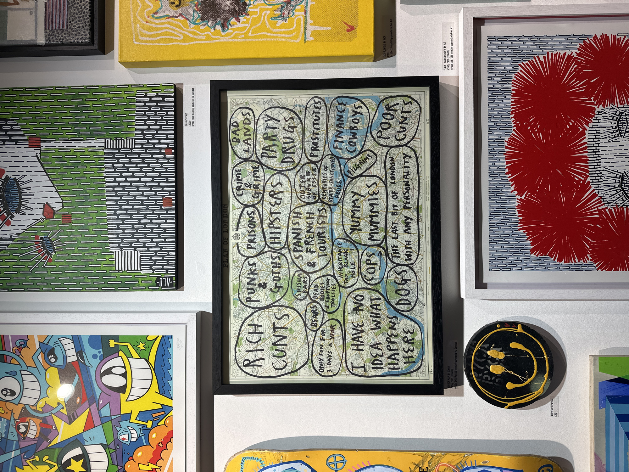 The image captures a section of an art gallery wall, displaying multiple contemporary artworks in various styles. The central piece is a framed map of London, overlaid with numerous hand-drawn, irregular bubbles containing a satirical or critical commentary on different aspects of the city. These labels include phrases such as "RICH CUNTS," "PUNKS & GOTHS," "HIPSTERS," "PARTY DRUGS," "PROSTITUTES," "FINANCE COWBOYS," "POOR CUNTS," "YUMMY MUMMIES," and "I HAVE NO IDEA WHAT HAPPENS HERE," among others, signed "Edwin."
To the left of the map, a framed graphic artwork features a green background with white and black horizontal dashed lines, depicting part of a face with an eye. Above it, a yellow canvas with abstract markings is partially visible. To the right, another framed artwork showcases a blue and white striped background adorned with large, vibrant red, spiky circular forms. Below this, a small, circular black object with yellow splatters and a distorted smiley face design is mounted on the wall. At the bottom of the frame, a yellow skateboard deck with colorful cartoon illustrations is visible.
Pricing and artist information are displayed next to some pieces. The green artwork is "SOPHIA" by ALO, priced at £3000. The central map is "LONDON MAP" by Edwin, priced at £475/£625 (framed). The red artwork is "ACID FLOWERS" by Ryca (£150) and "LADYFLOWER CROWN" by ALO (£350/£600 framed). The round black piece is "POPPERS" by Ryca, £50. Payment options via "Own Art" are also noted, indicating a retail art setting. The overall impression is a vibrant display of urban-themed and graphic art.