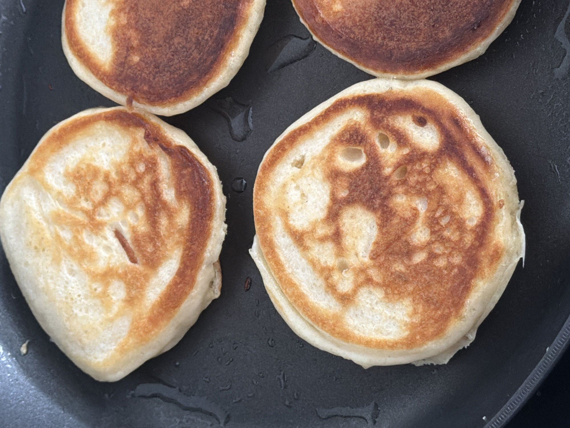 This is a close-up, overhead shot of four golden-brown pancakes cooking in a dark frying pan. The pancakes are round and puffed up, with visible air bubbles on their surfaces. The pan has a non-stick surface and some oil is visible around the edges of the pancakes. There are no people or other objects in the frame. The lighting is natural and appears to be from above. This scene depicts a common culinary activity, likely taking place in a kitchen in Finestrat, Spain, given the request.