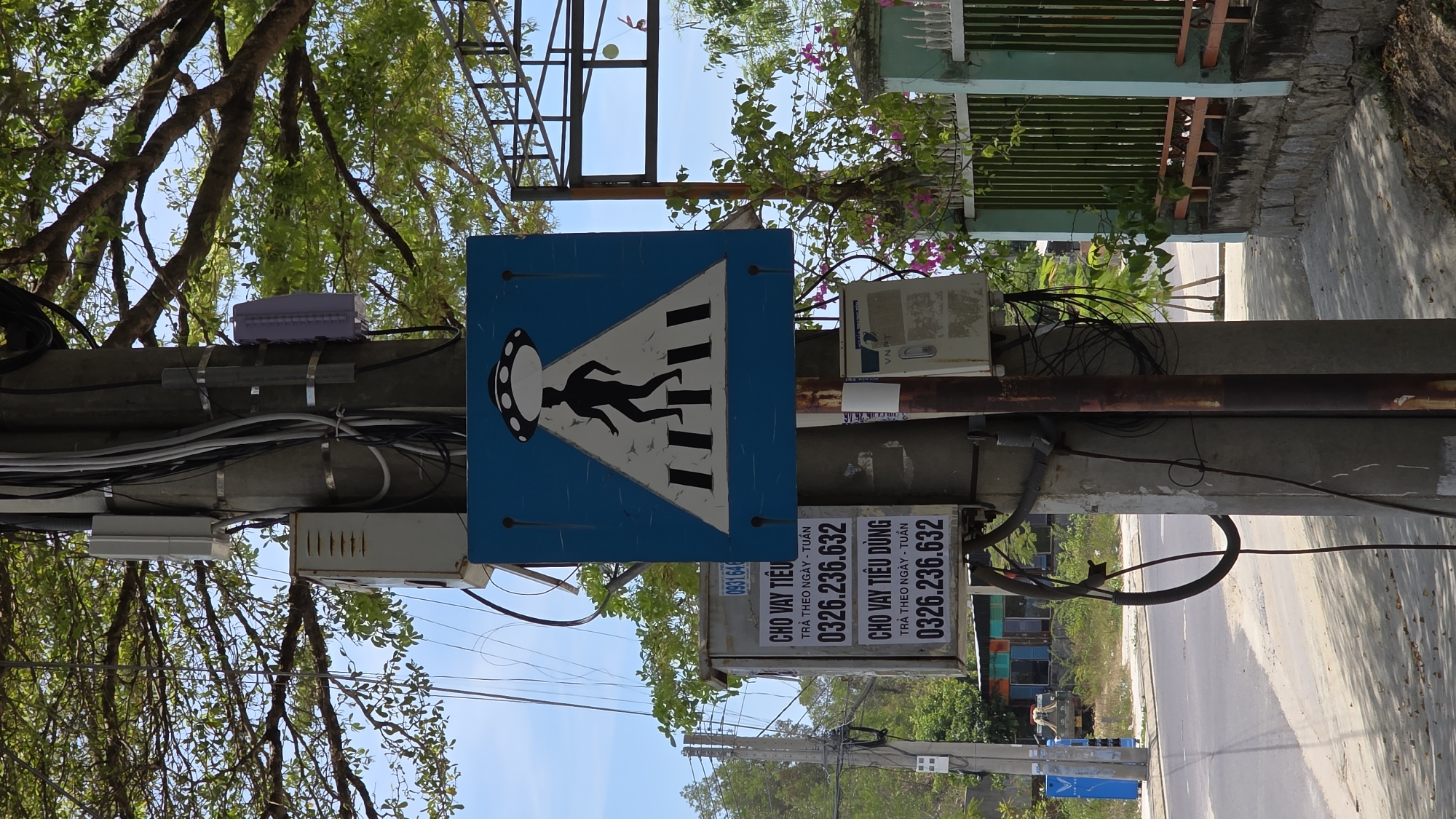 The image captures a utility pole in Nha Trang, Vietnam, displaying an unusual pedestrian crossing sign with a person walking towards a UFO, set against a blue triangular background with zebra crossing stripes. The pole also features advertisements for "CHO VAY TIÊU DÙNG" (consumer loans) and the phone number 0326.236.632, indicating daily/weekly payment plans. Additionally, there's a small VNPT (Vietnam Posts and Telecommunications Group) sign and various electrical boxes and wires attached to the pole. Vegetation is visible in the background, with trees partially obscuring the sky. The street and a building are seen to the right of the pole.