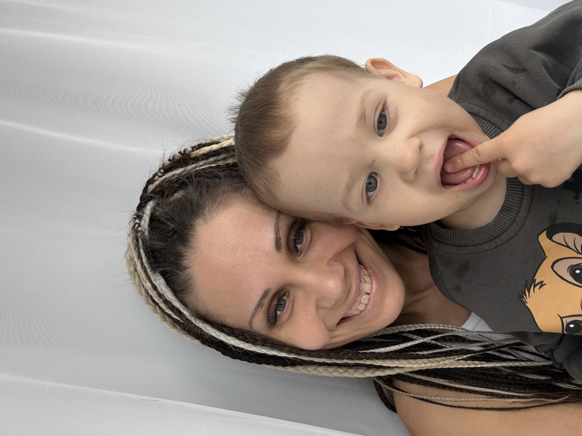 A close-up, portrait-style photograph captures a joyful moment between an adult woman and a young child, likely a mother and son. The woman, positioned on the left, smiles warmly at the camera. She has light-colored eyes and long, distinctive braided hair (dreadlocks) in mixed shades of dark brown, light brown, and white/grey. The child, a young boy with light brown hair and blue eyes, rests his head affectionately against the woman's, facing playfully to the right. He makes a comical expression, with a finger in his open mouth. He is wearing a dark grey long-sleeved shirt featuring a graphic of Simba from Disney's The Lion King. Both subjects exhibit clear emotions of happiness and playfulness. The background is a plain, light-colored fabric, possibly a white sheet or curtain, with subtle folds and textures, suggesting a simple indoor setting. The soft, even lighting creates an intimate and cheerful atmosphere, consistent with a casual moment in a home, potentially in Rusanovo, Russia.