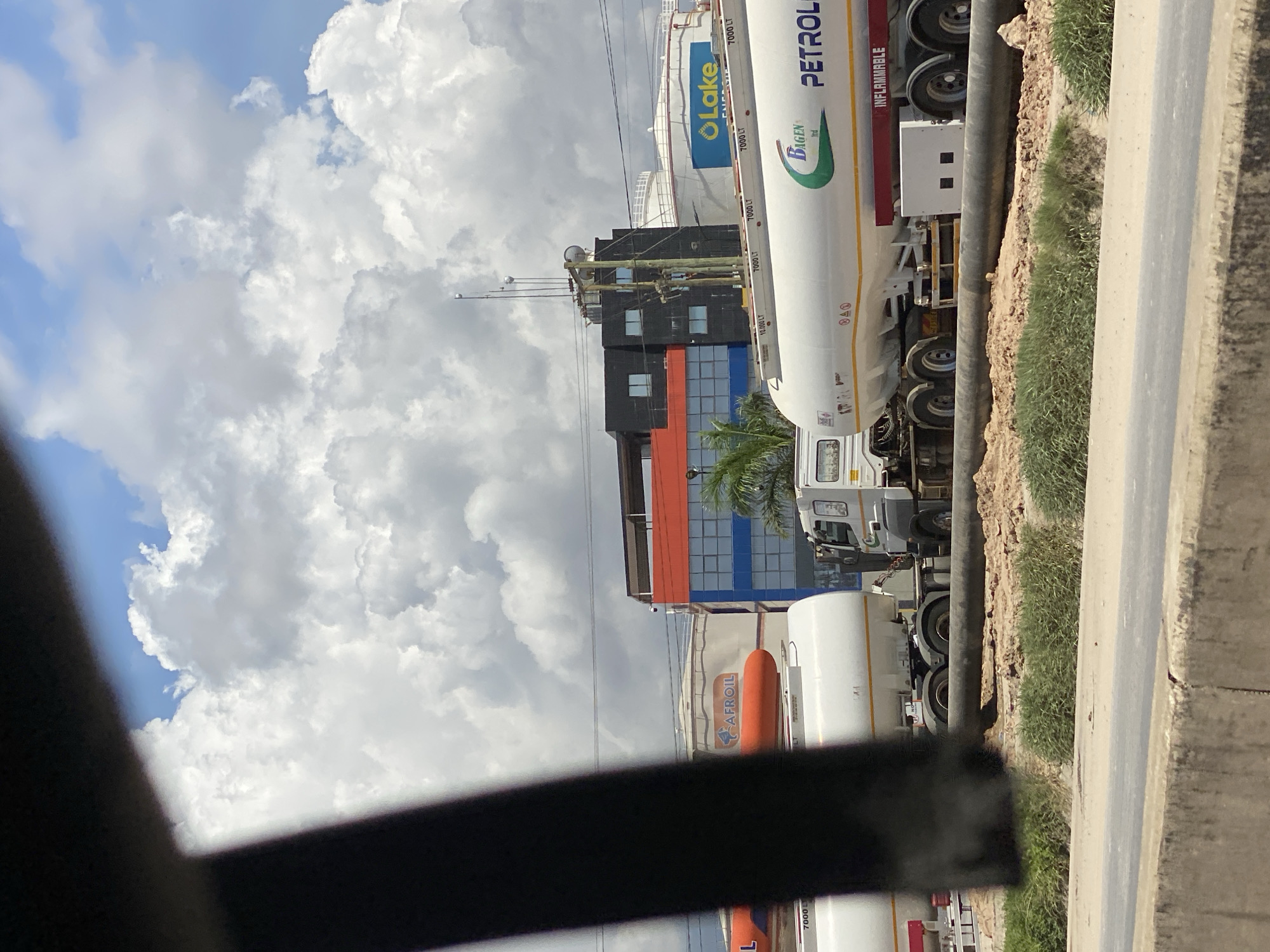The image shows a street scene in Dar es-Salaam, Tanzania. In the foreground, there is a black metal structure partially obscuring the view. Along the roadside, a series of oil tanker trucks are parked, some bearing the logos of "Lake Energy" and "AFROIL" and "PETROL". The trucks are white with either blue or orange accents. Behind the trucks, there is a building with a black, red, and blue facade, indicating a corporate or industrial area. A palm tree partially obscures the building. The sky is partly cloudy, with a mix of blue sky and large white clouds. On the right, there is a concrete curb and a patch of green grass or vegetation.