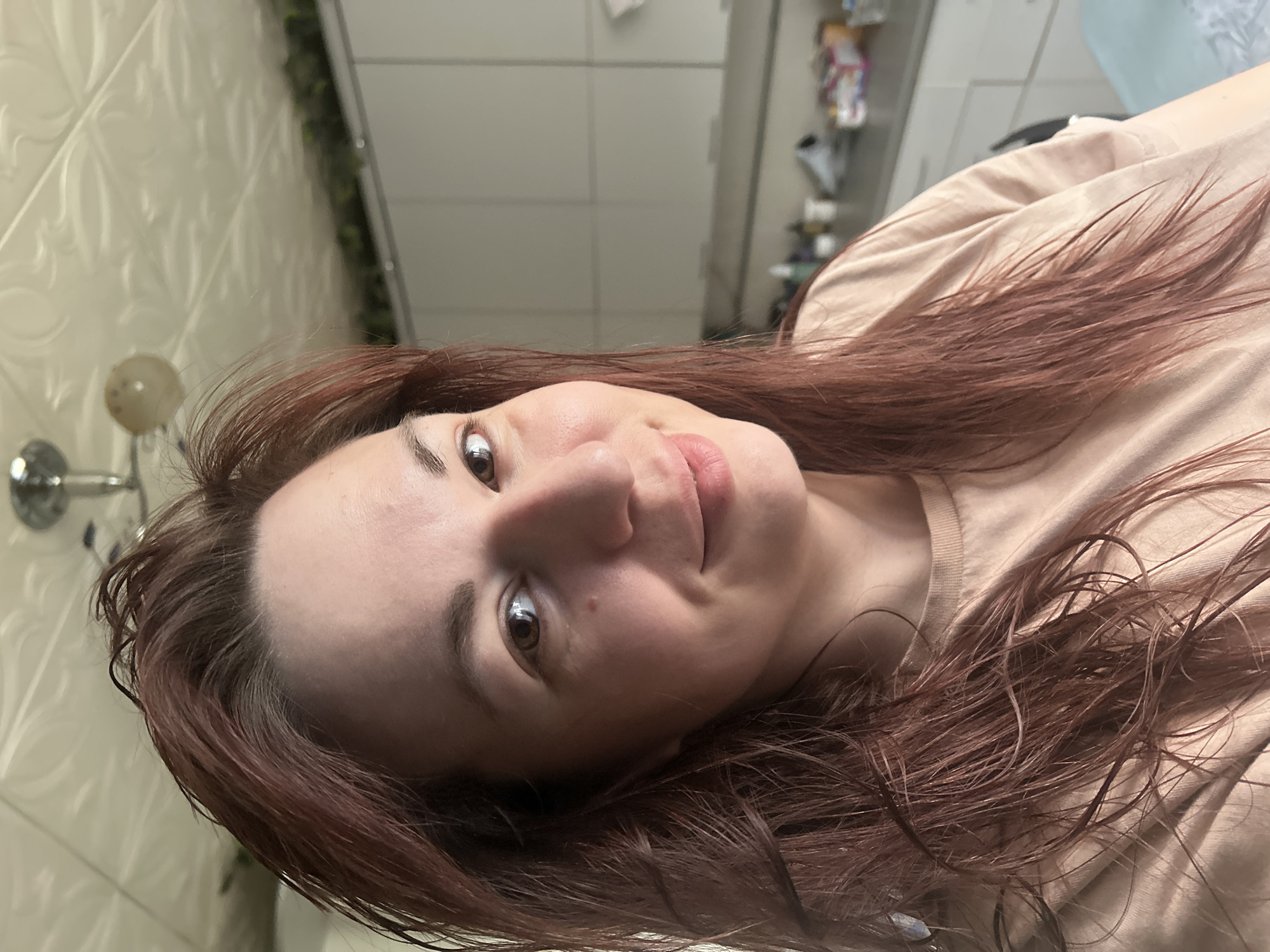 A woman is looking at the camera, with wet reddish-brown hair, wearing a beige top. The background shows a bathroom with patterned white wall tiles, a wall-mounted light fixture, and a shelf with miscellaneous items. There is no verifiable evidence the location is in Фоки, Russia.