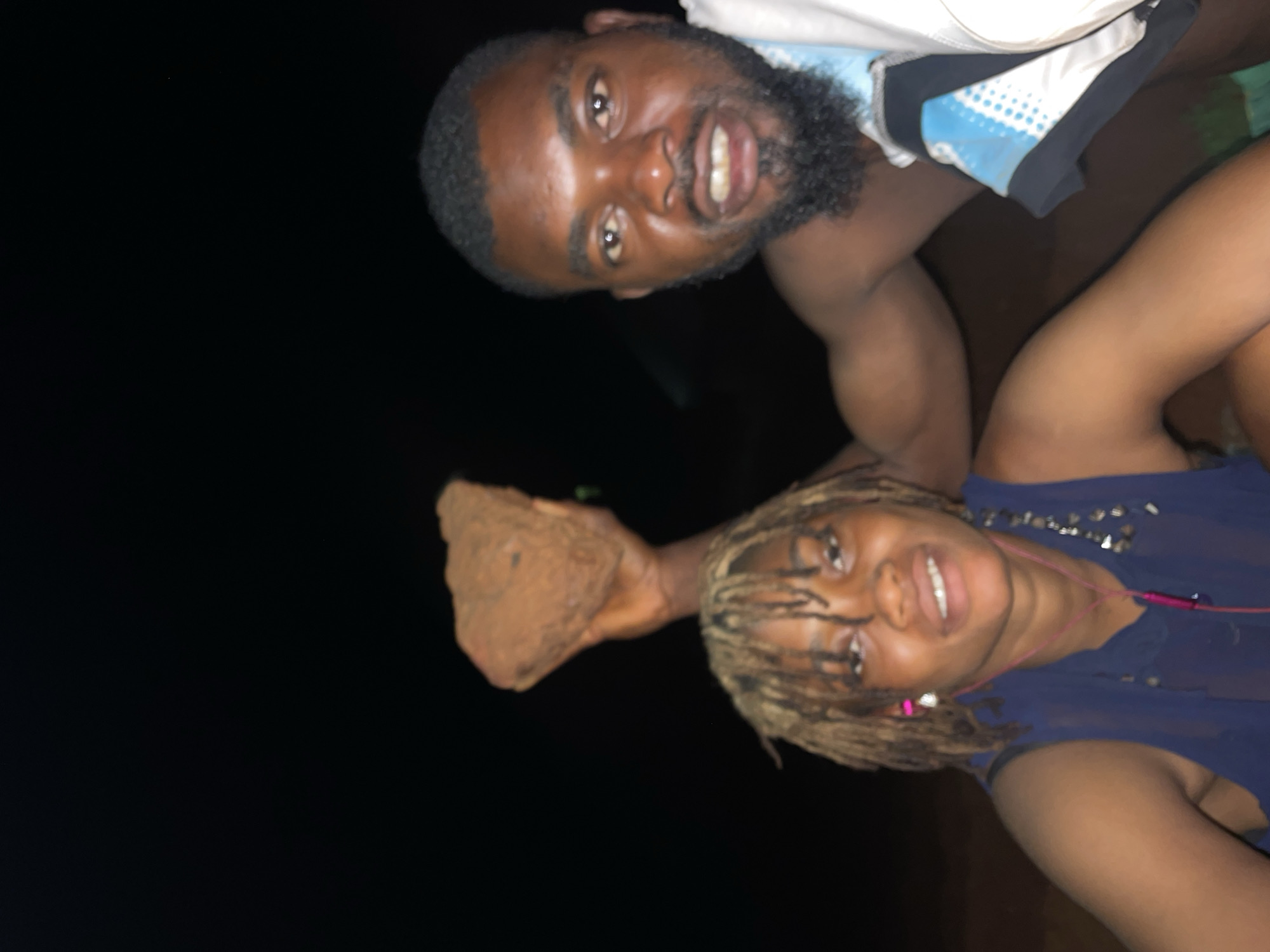 A nighttime photograph depicts two individuals outdoors. A male with dark hair and a beard wears a light-colored shirt with blue detailing. He is positioned above and to the right of a female with light brown dreadlocks. The female wears a dark blue top and holds a clod of dirt in her hand. The background is dark, suggesting a low-light environment. No specific location is discernible beyond the general environment.