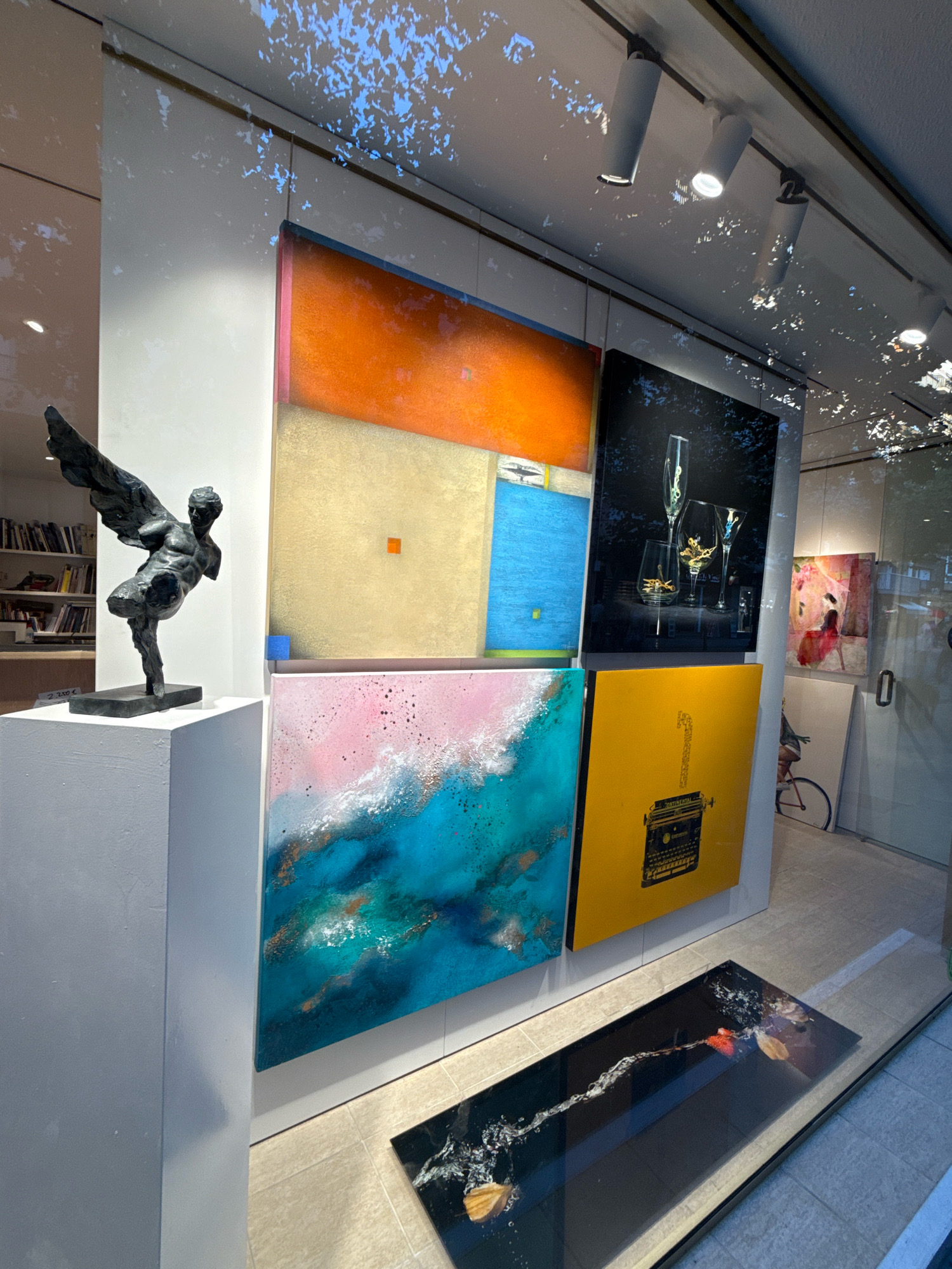 The image shows an art gallery display window in Blanes, Spain. The window features a variety of artwork, including paintings and a sculpture. In the foreground, a dark bronze sculpture of a winged figure stands on a white pedestal. To the right of the sculpture, four framed paintings are displayed. The top left painting has a gradient of orange and gold. The top right painting features champagne glasses with golden objects inside them. The bottom left painting is a mix of pink and turquoise with white and gold accents. The bottom right painting is bright yellow with a black silhouette of a typewriter. On the floor in front of the paintings is a rectangular artwork on a dark background, depicting a fish in water with leaves. In the background, shelves filled with books are visible. Through the window, a glimpse of the street and foliage can be seen. The lighting suggests it is daytime.