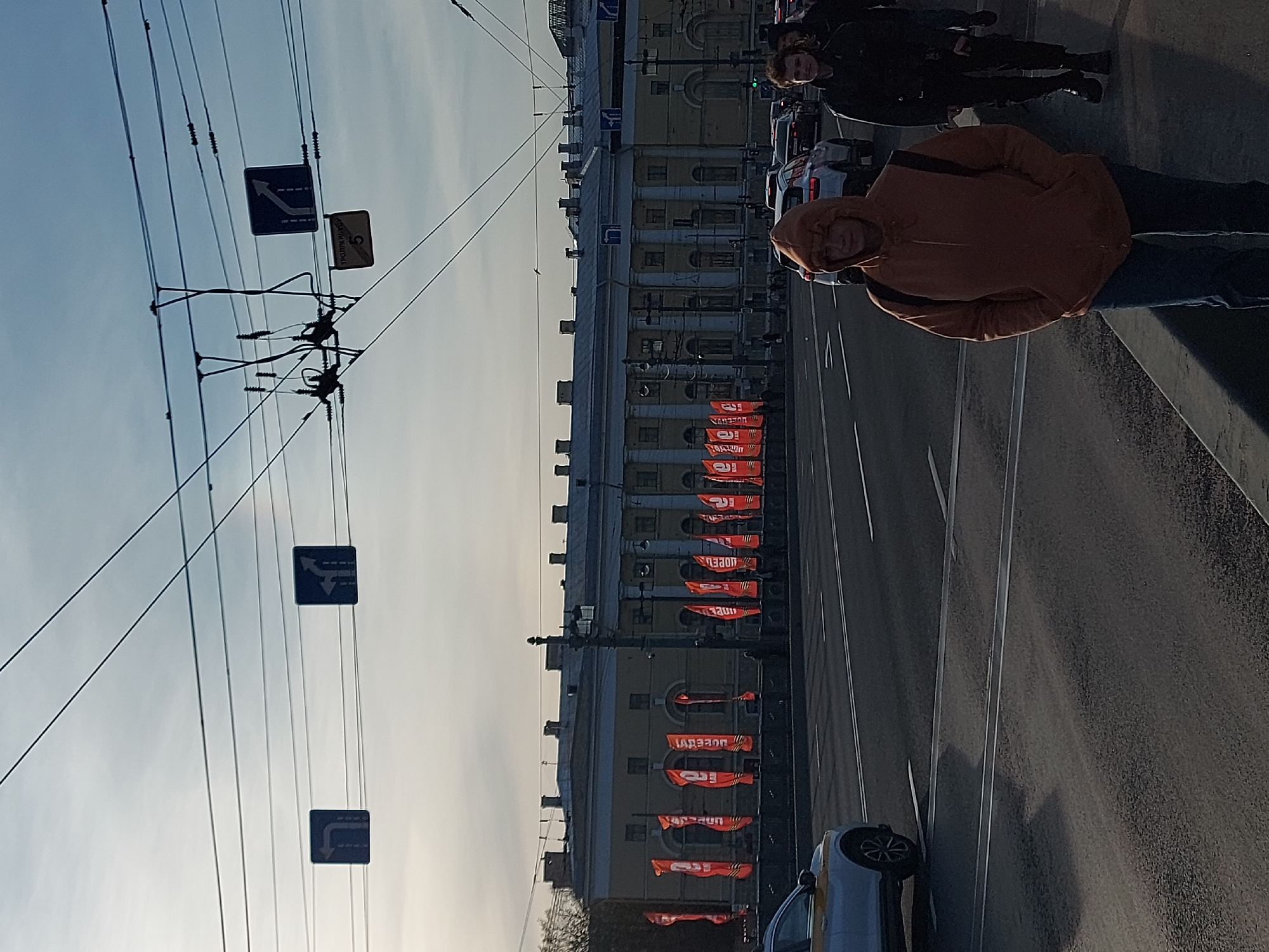 The image depicts a street scene in Saint Petersburg, Russia, likely along Nevsky Prospekt. The foreground contains two people, one wearing a brown hooded jacket and another wearing a black jacket. Both are standing on the sidewalk, close to the roadway. Cars and a building line the street. The building has orange flags, likely a display for Victory Day, with the word "Победа" (Victory) visible on some of them. Above the street are power lines and traffic signs.