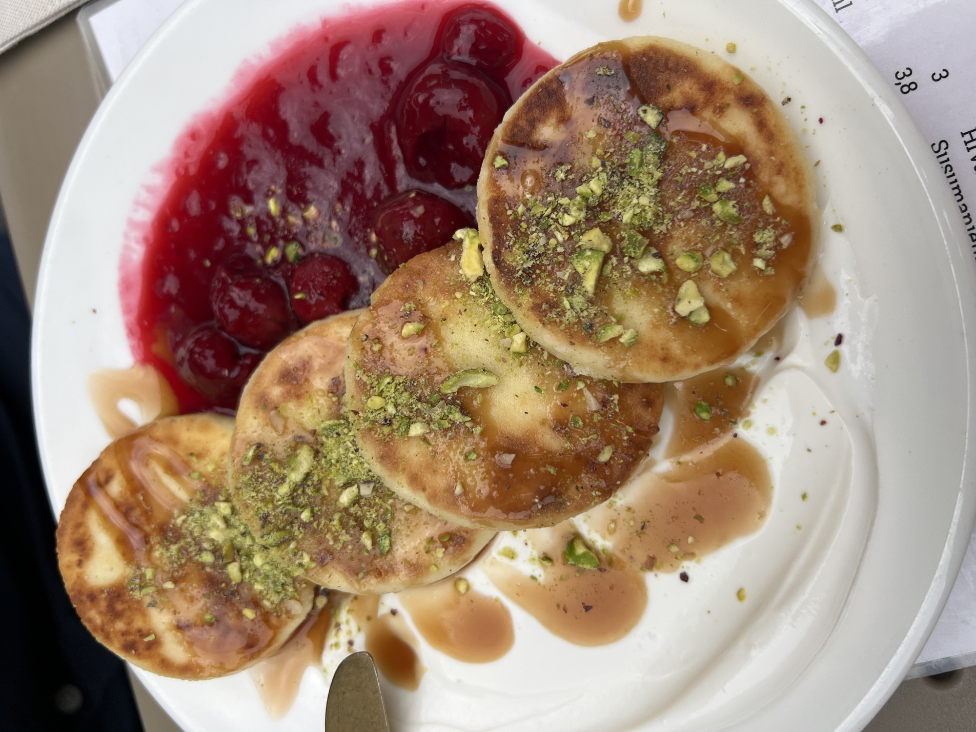 The image shows a white plate with several syrniki (small cheese pancakes) garnished with chopped pistachios, accompanied by a generous serving of cherry sauce and a dollop of white cream. The syrniki are golden brown and drizzled with a light syrup. The dish appears to be photographed at an outdoor restaurant. Text in an adjacent document partially reads "Susimania" and numbers appear, possibly related to pricing in EUR. The precise location is within Podgorica, Montenegro.