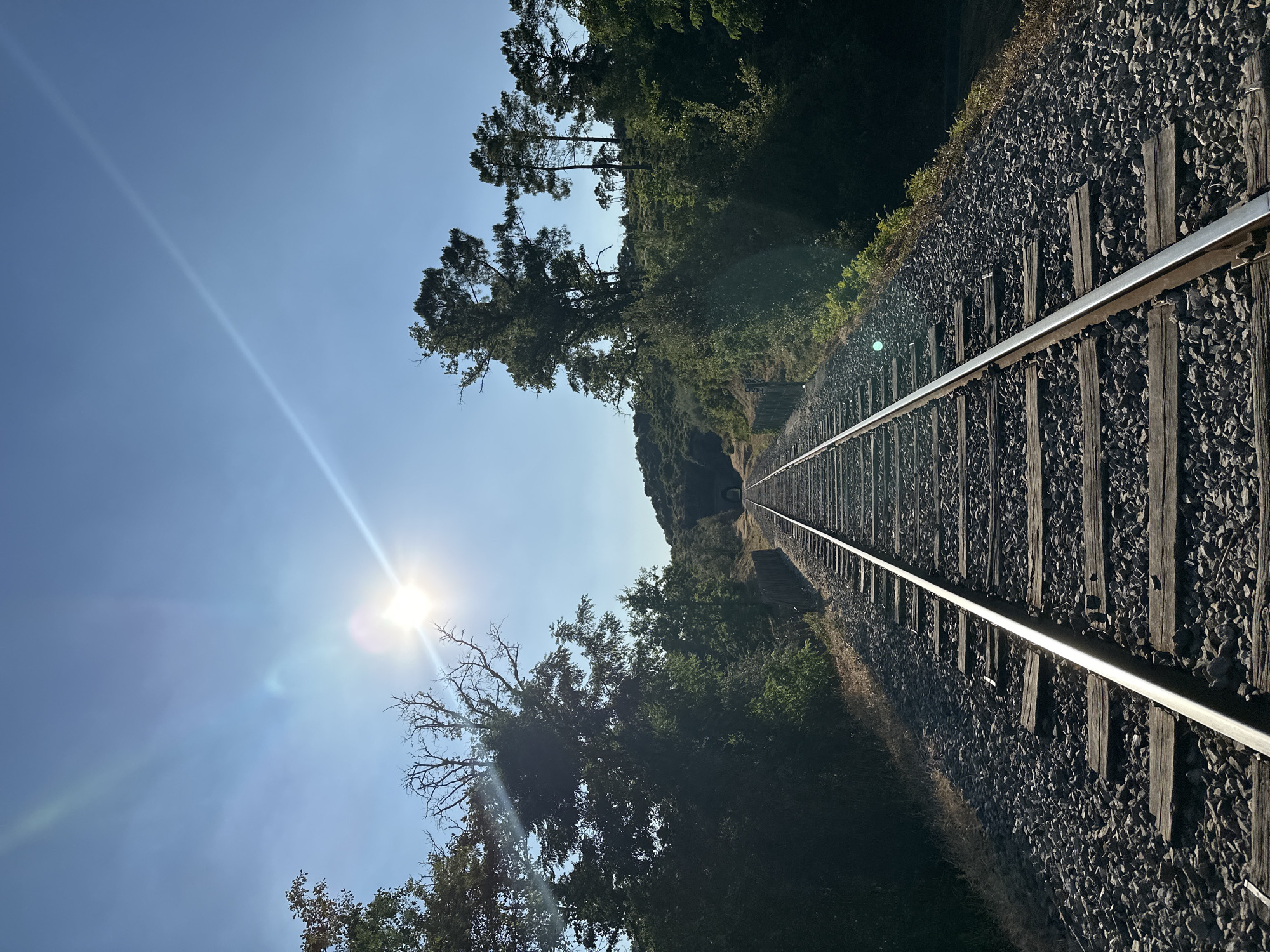 The image shows a railway track leading towards a tunnel in a rural area near Générargues, France. The sun is shining brightly, creating lens flare in the sky and illuminating the scene. Trees and vegetation line the sides of the tracks, which are laid on a bed of ballast. The sky is clear blue. The scene is peaceful and natural, with no people or animals visible.