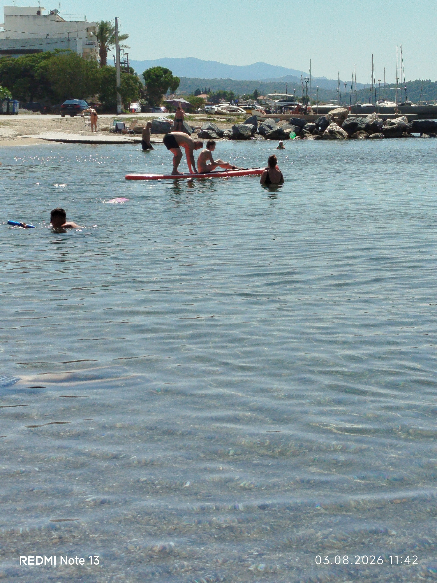 The image depicts a sunny day at a beach in Sithonia Municipal Unit, Greece, with a clear, shimmering sea in the foreground. In the water, several individuals are visible. A person on a red paddleboard is in the center, with another person appearing to be in the water near them. To the left, a person is swimming, holding a blue flotation device. Further out in the water, a person is partially submerged. The background reveals a shoreline with rocks, vegetation, and buildings, including a multi-story white structure. Boats are docked in a harbor further along the coast, and mountains are visible in the distance under a clear sky. The water is shallow in the foreground, with the seabed visible through the translucent water. The scene captures a typical summer day at a Greek coastal location.