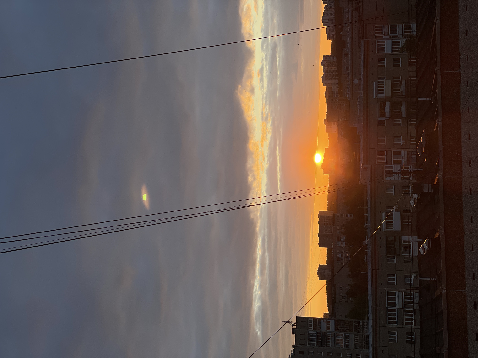 This is a vertically oriented, outdoor photograph taken in Izhevsk, Russia, depicting a city skyline at sunset. The sun, a bright, intense orb, is low on the horizon, casting a warm, orange glow across the sky and illuminating a band of clouds with golden light. The sky transitions from a pale orange near the sun to a soft gray and then a darker gray towards the left side of the frame.
In the foreground, three dark power lines stretch horizontally across the image, obscuring portions of the sky and the cityscape. The cityscape itself consists of a series of multi-story apartment buildings, typical of Soviet-era architecture, that recede into the distance. The buildings are primarily rendered in muted tones of gray and beige, with visible windows and balconies. Some trees are visible behind the buildings, appearing as dark silhouettes against the bright sky. The overall impression is one of urban life juxtaposed with the natural beauty of a sunset. The lighting suggests it is evening. There is no visible text in the image.