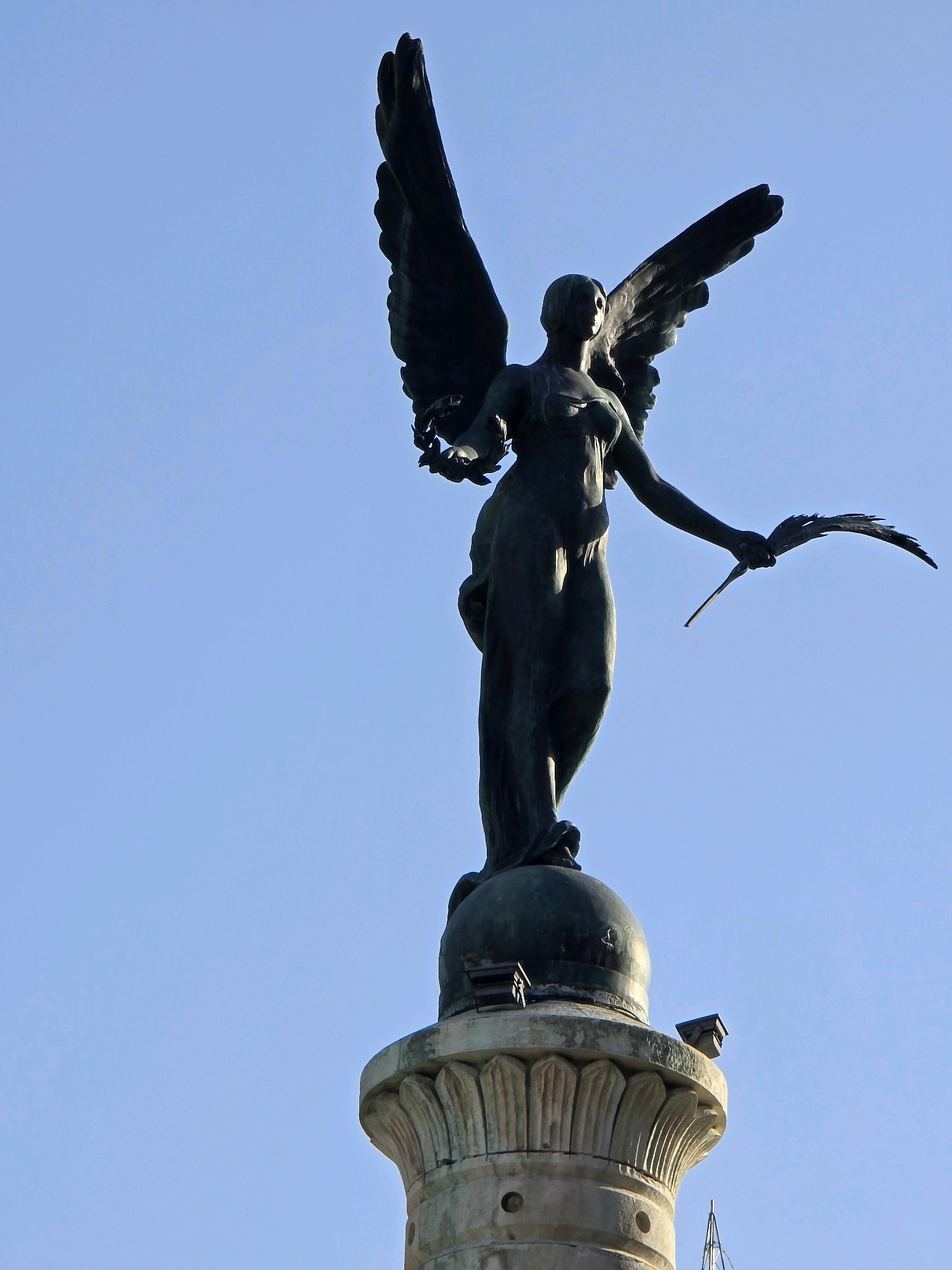 The image is a low-angle shot of a bronze statue of a winged figure, likely a representation of victory or an angel, standing atop a classical-style pedestal. The figure is depicted in mid-stride, with one arm outstretched holding a laurel wreath and the other arm bent, holding a palm frond. The statue has large, outstretched wings, and is dressed in flowing robes. The pedestal is made of stone and features fluted details. The background is a clear blue sky. The statue is located in Plaza de las Batallas, Jaén, Spain, and is known as the Monumento a la Batalla de las Navas de Tolosa.
