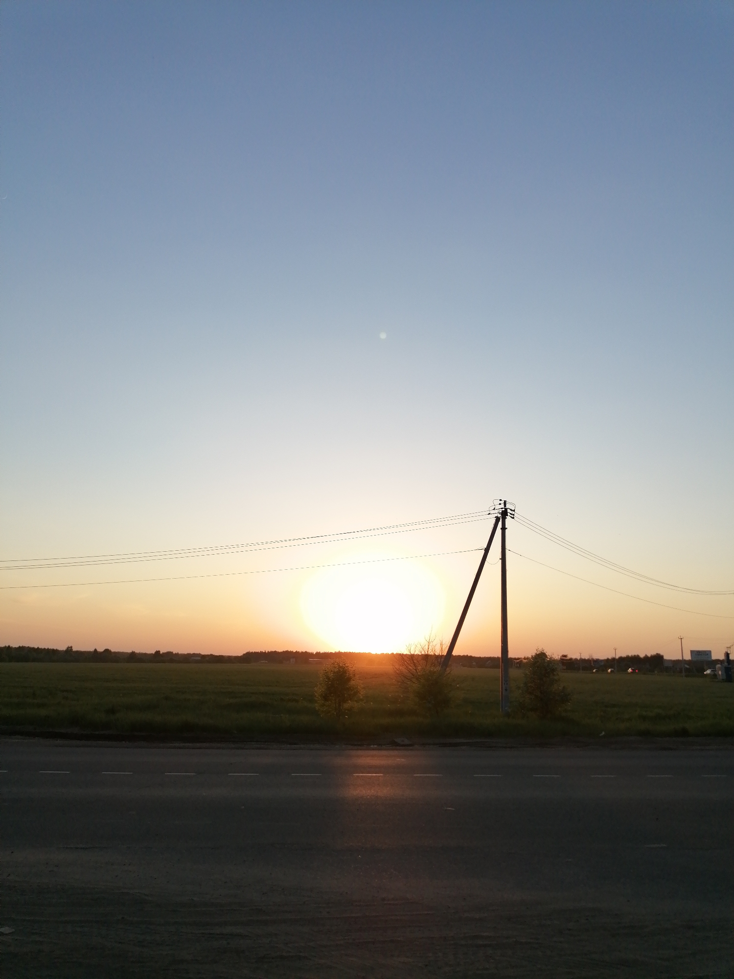 The image shows a landscape at sunset. The sun is low on the horizon, creating a bright, orange glow. A grassy field stretches out in the middle ground. A utility pole stands to the right of the sun, with power lines extending across the scene. There are a few small trees scattered around the pole. In the foreground, there is a paved road with some sunlight reflecting off its surface. The sky is a gradient of blue and orange, fading from light blue at the top to orange near the horizon.