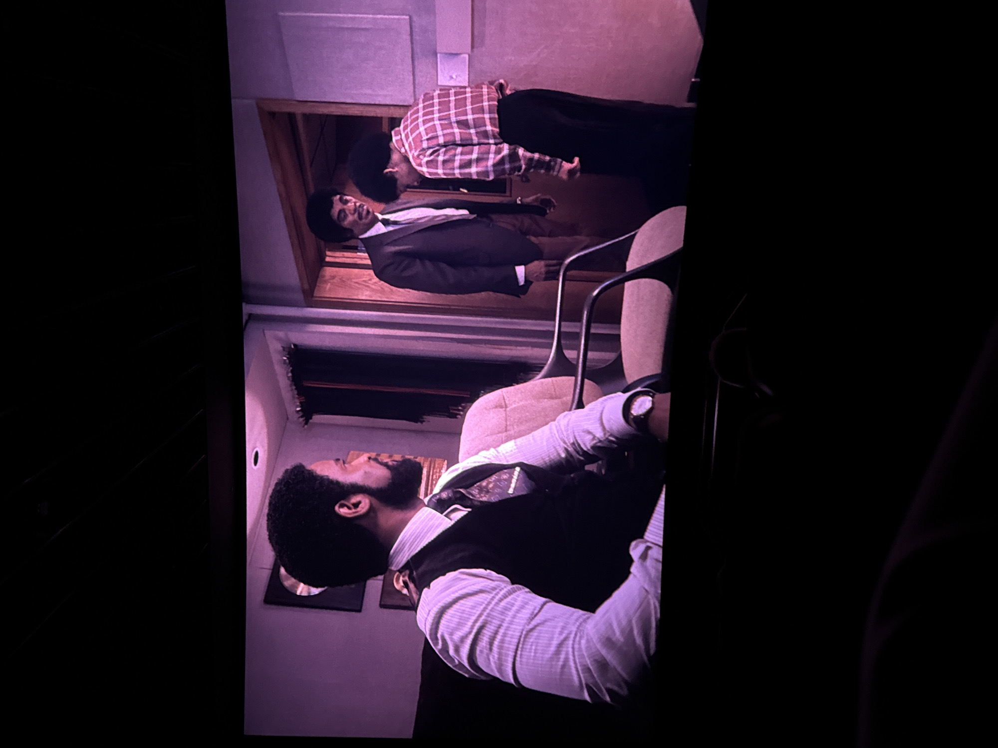 Here is a detailed description of the image:
The image shows three Black men in an indoor setting with purplish lighting. The scene appears to be a set from a movie or television show.
One man is seated in a chair in the foreground, wearing a white shirt, a dark vest, and has a full beard and afro. He is looking to the left of the frame.
In the background, two men are visible through a doorway. One man is wearing a dark suit and the other a red and white checkered shirt and dark pants. It appears the two men are either in conversation or engaged in some sort of confrontation.
The environment consists of walls and furniture, suggesting an office or an interior space. The color palette is dominated by purple hues. There is no information available in the image about the location being in Sofia, Bulgaria.
