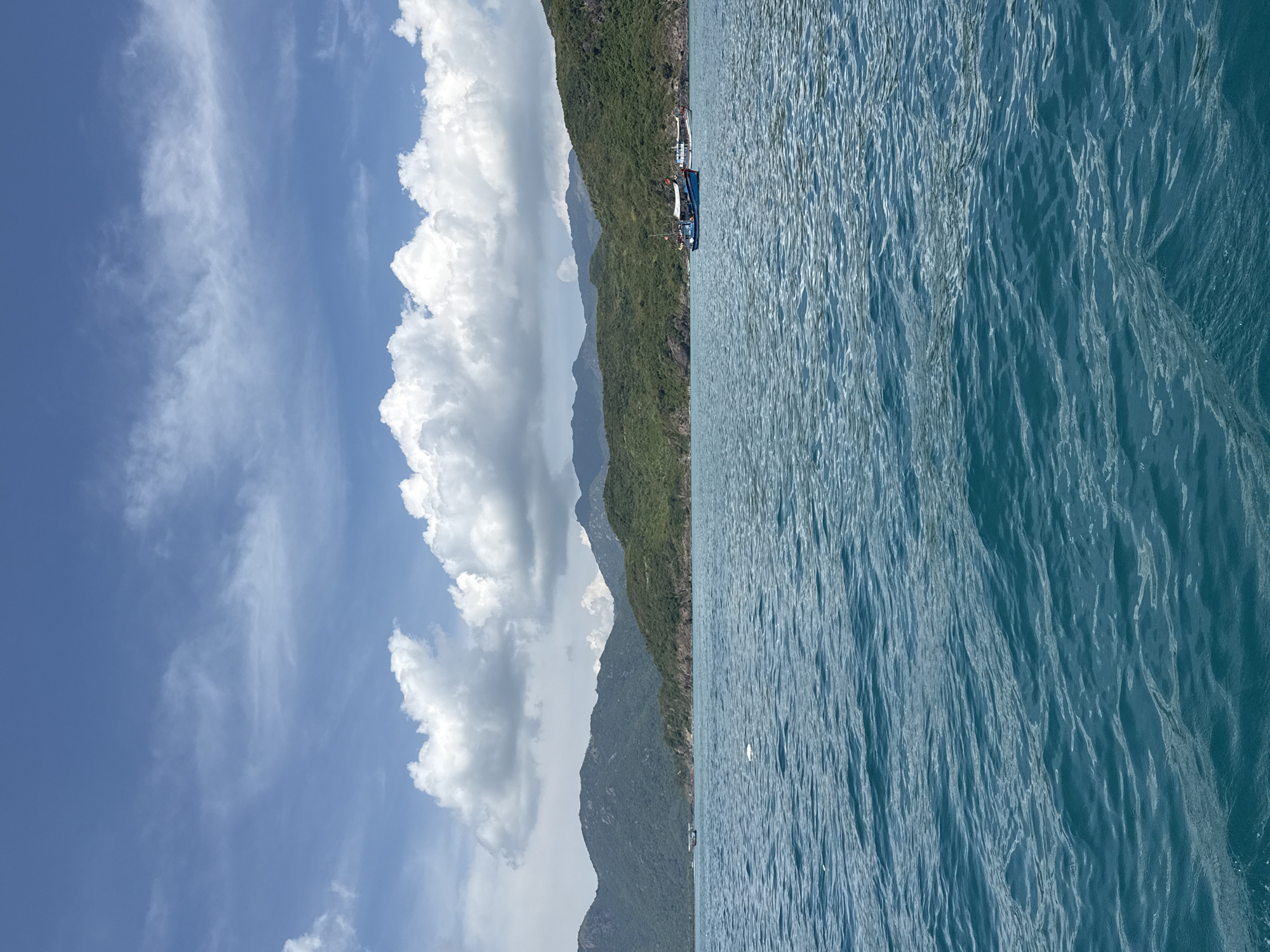 The image depicts a coastal scene in Nha Trang Ward, Vietnam. The sky is partly cloudy, with large cumulus clouds adjacent to a mountainous coastline covered in dense green vegetation. Several boats are docked along the coastline. The ocean occupies a significant portion of the image, displaying textured, slightly choppy water. There are no visible people in the image.