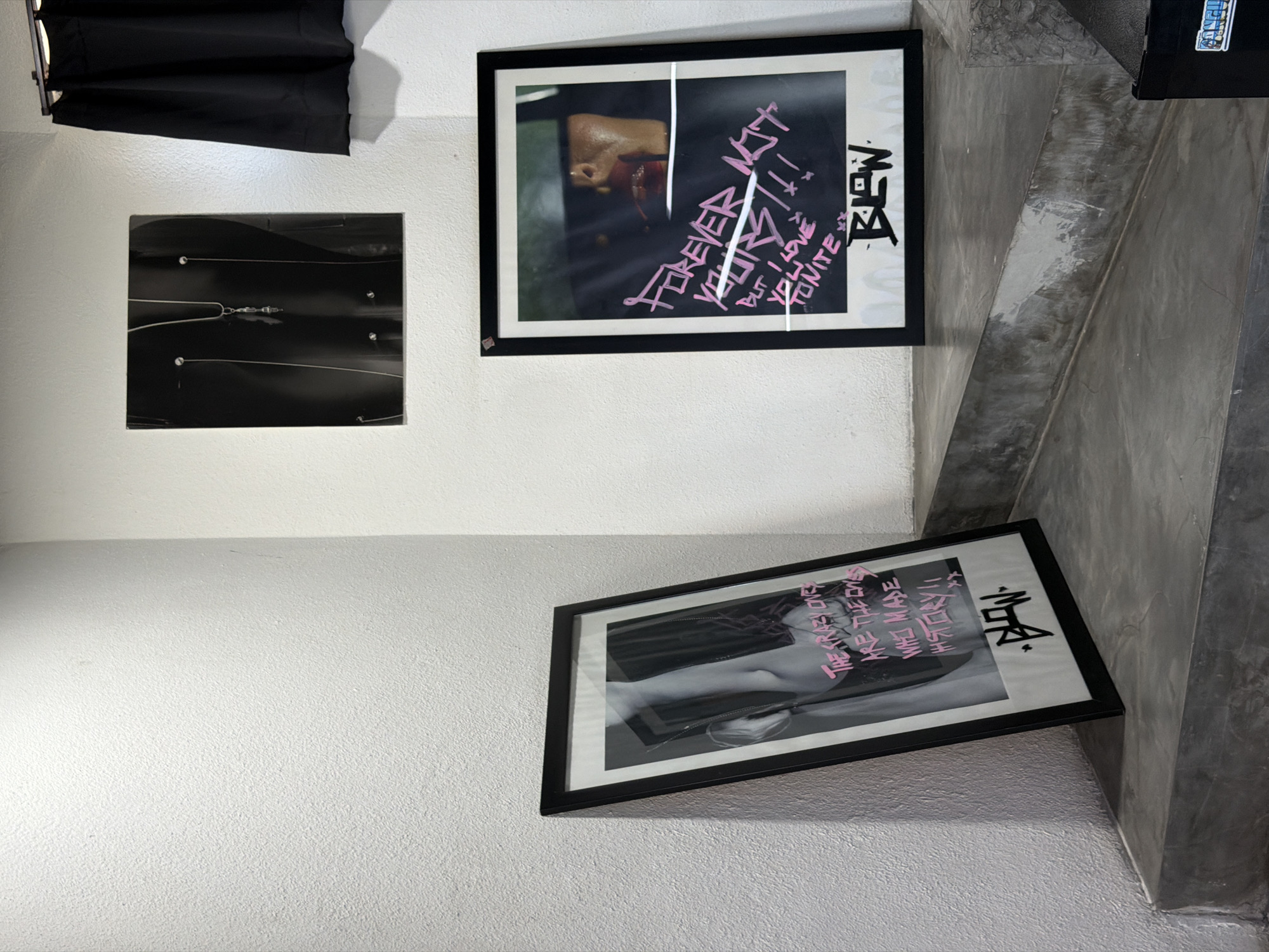 The image depicts an indoor corner with white textured walls and grey concrete steps. On the wall, a framed black and white photograph with pink text is displayed. Below this photograph, another framed artwork with a dark background and pink graffiti-style text is leaning against the wall on the top step. To the right of this artwork, a black curtain is visible. The scene suggests a retail or gallery space in North Kuta, Indonesia.