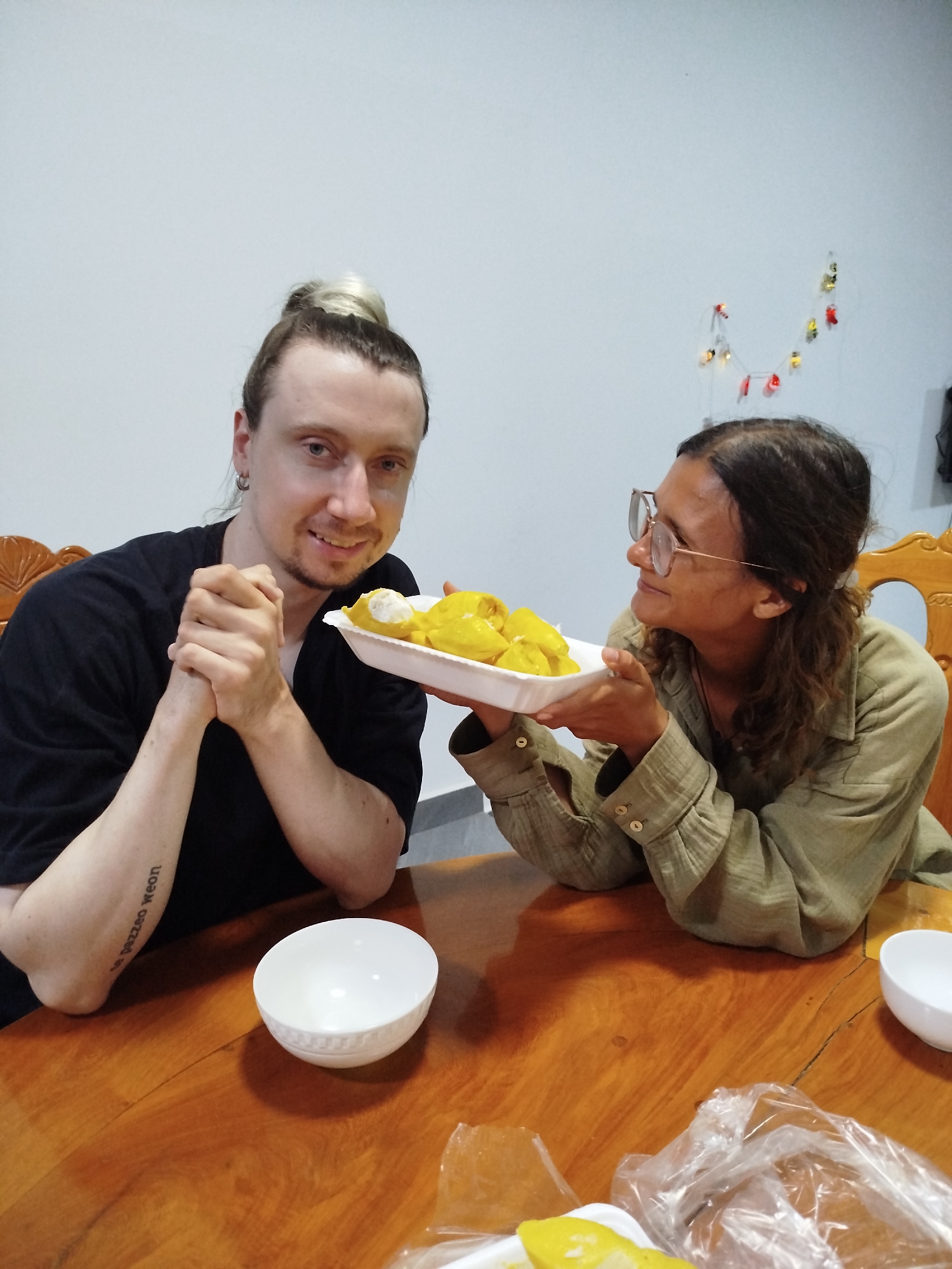 Two individuals are seated at a wooden table indoors. On the left, a fair-skinned individual with hair in a bun wears a black shirt and has a tattoo on their left forearm. They have clasped their hands together. On the right, another individual wearing glasses and a light green button-down shirt holds a white styrofoam tray filled with yellow food items, possibly durian. They are gazing toward the person on the left. In front of each person is a white bowl. Plastic bags and additional durian pieces are visible on the table. In the background, a light-colored wall is adorned with small decorative lights. The scene is likely set in Da Lat, Vietnam, based on the presence of durian, a fruit commonly associated with Southeast Asia.
