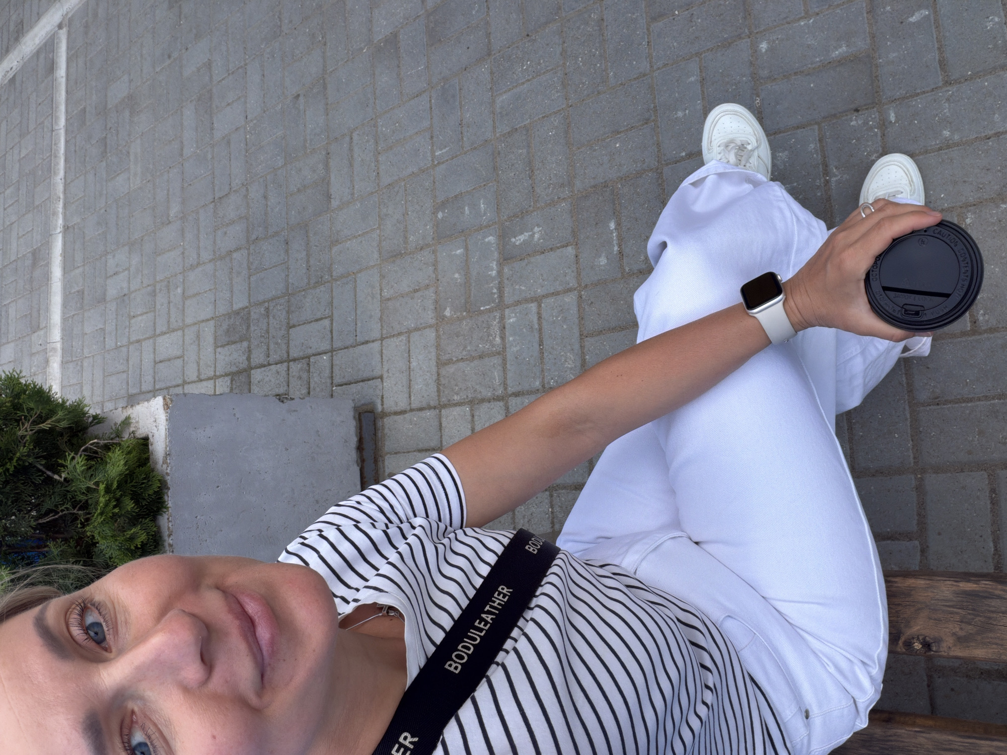 A woman in a black and white striped shirt and white pants and shoes sits on a bench, holding a black lid to a coffee cup. She is wearing a white smartwatch. The ground is paved with gray blocks. The photo appears to be taken in Chaikovsky, Russia.