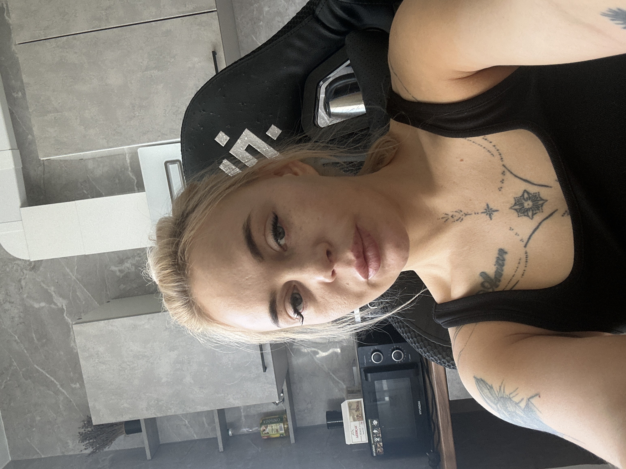 A medium, slightly-angled shot presents a fair-skinned woman with blonde hair pulled back. She is wearing a black tank top and seated in a black gaming chair. A pattern of tattoos are visible on her chest and upper right arm. The background reveals a kitchen setting with grey cabinets and countertops. A microwave, labeled "Hyundai", sits on the counter behind her. The location is suggested to be Высокий, Russia. There is no information to confirm or deny the location.