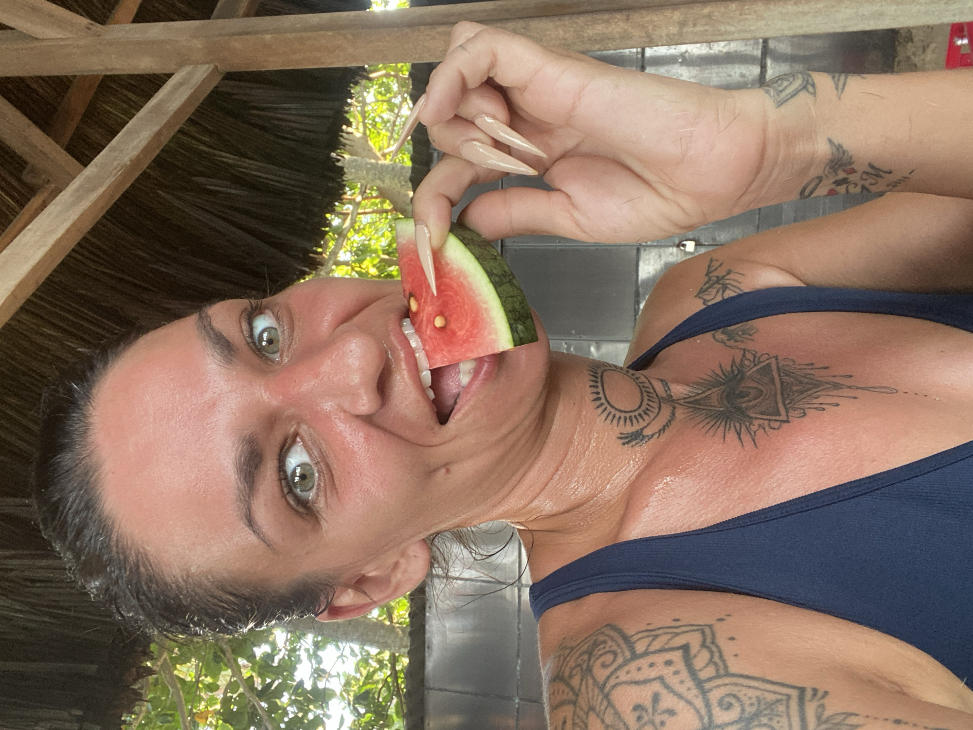 In Mũi Né, Vietnam, a woman with short dark hair pulled back and a blue tank top is captured in a close-up selfie. She has green eyes and her face is flushed, suggesting sun exposure. She is holding a slice of watermelon to her mouth, which she is about to eat. The background shows a thatched roof and wooden structure, indicative of local architecture, with lush green foliage visible behind her. The woman has several tattoos, including an intricate mandala on her left shoulder and arm, an eye symbol on her neck, and a smaller tattoo on her right wrist. Her fingernails are long and pointed. The image appears to be taken outdoors, in what seems to be a relaxed, possibly tropical setting.