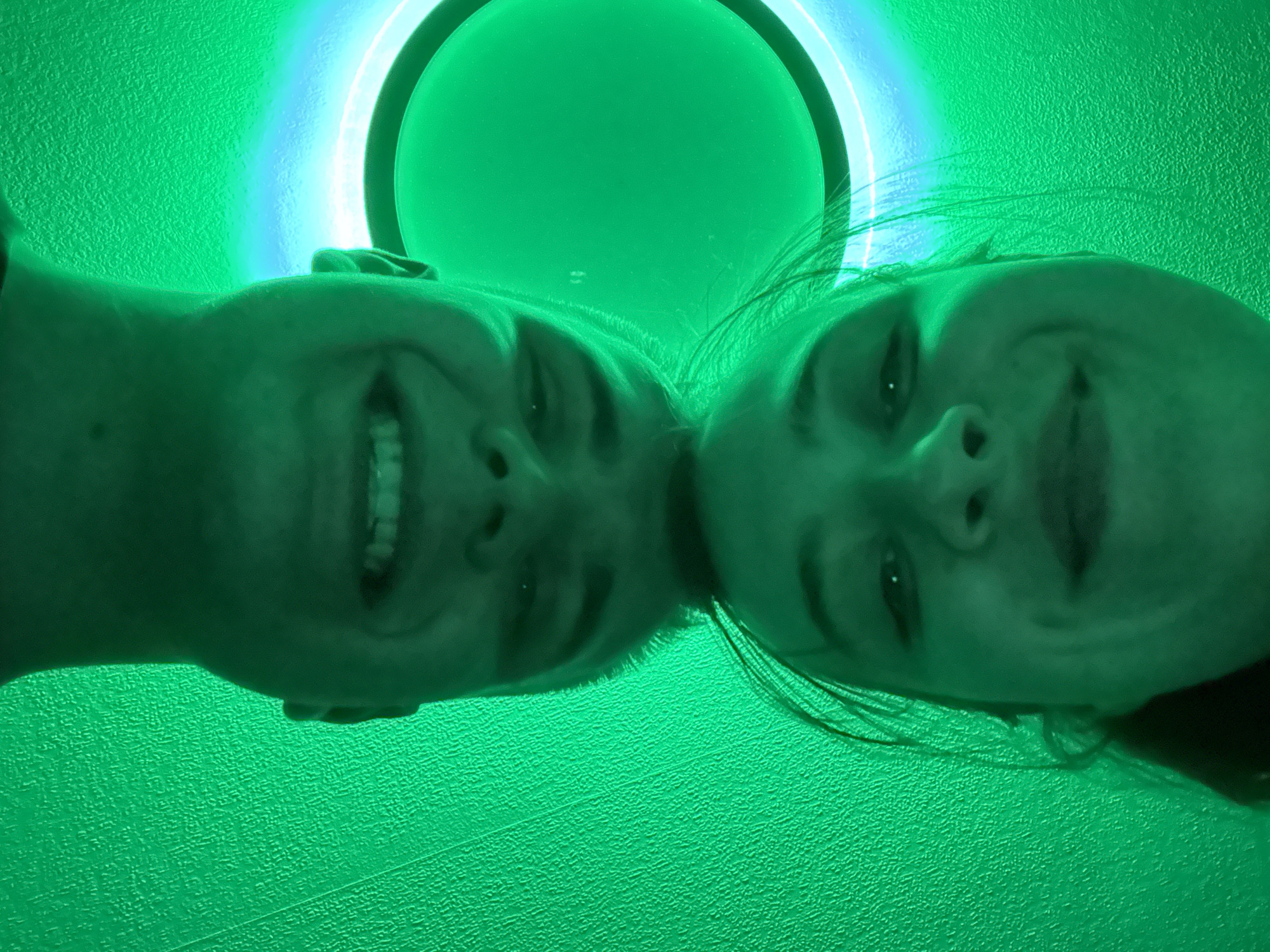 The image presents a low-angle, close-up perspective of two individuals, likely adults, looking downwards into the camera. Their faces fill the lower half of the frame, with the tops of their heads positioned closely together, almost touching. Both subjects are smiling broadly, conveying a sense of happiness or playfulness.
The entire scene is dominated by a strong green light, which casts a vibrant hue across their skin tones, hair, and the surrounding environment. Behind the individuals, a circular light fixture is prominently visible, acting as the primary light source. This fixture emits a bright green glow towards its center and a distinct blue light along its outer ring, creating a halo effect. The background appears to be a uniformly textured surface, possibly a wall or ceiling, completely bathed in the same intense green light. The artificial and monochromatic lighting suggests an indoor setting, such as a specialized room, a photo booth, or an entertainment venue designed with specific ambient lighting. No text is visible in the image. The time of day and external weather conditions cannot be determined.